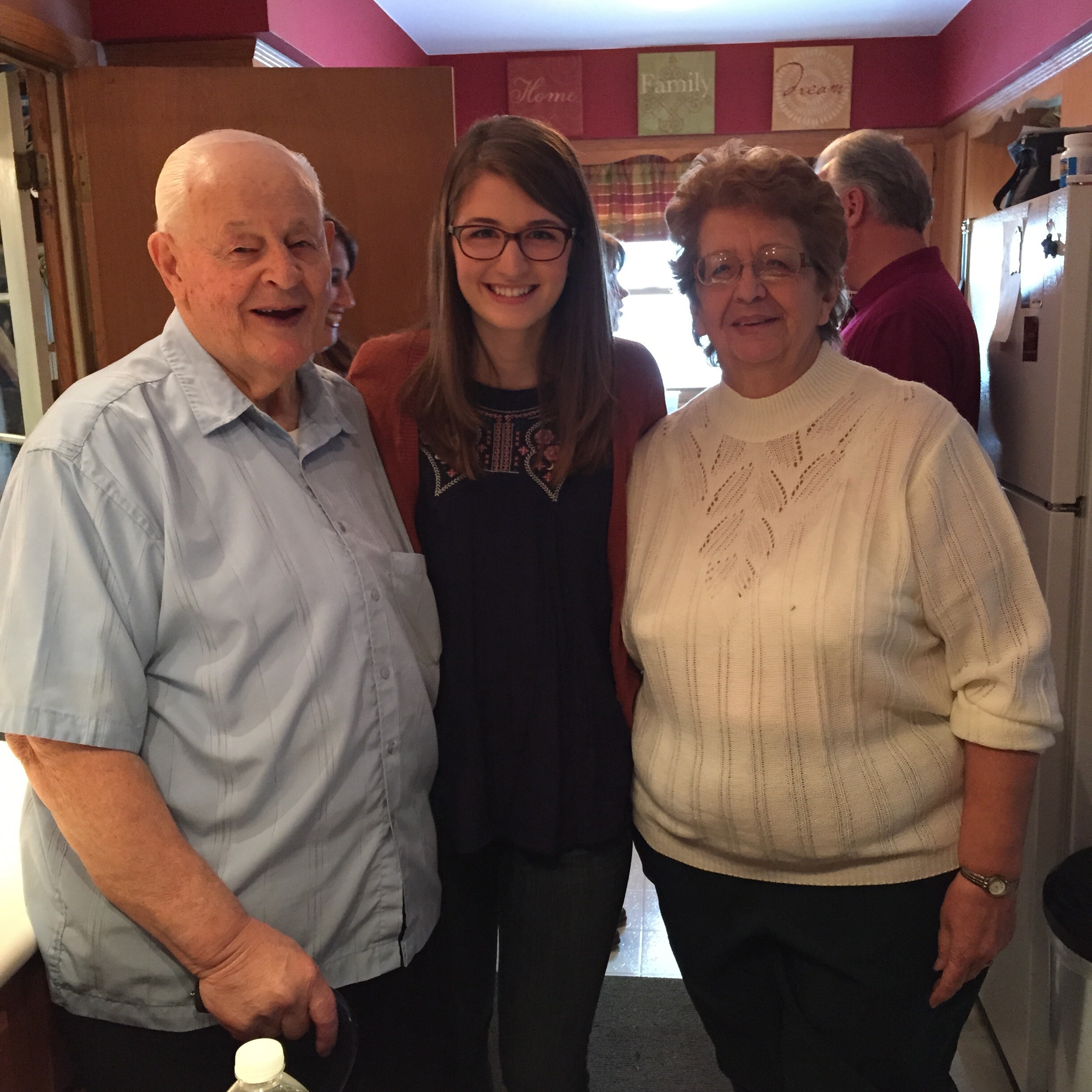 Three generations posing together in a kitchen: an elderly man on the left, a young woman with glasses in the center, and an older woman in a white sweater on the right, all smiling.