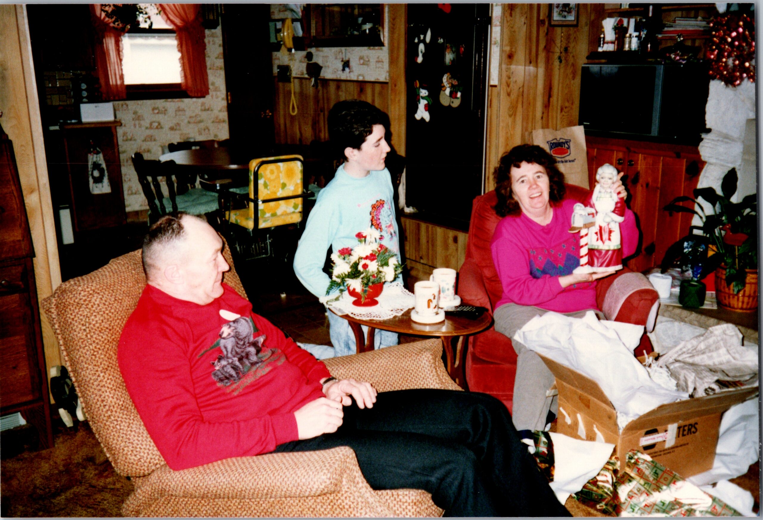 Family gathering in a cozy, wood-paneled living room as they open Christmas gifts; a woman in pink holds a Santa figurine centerpiece at the sofa.