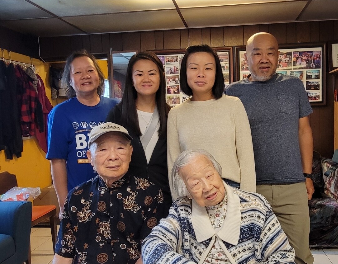 Eight people posing in a cozy room: two seniors in front, four adults behind them, and two more standing in back; framed photos on the wall behind.