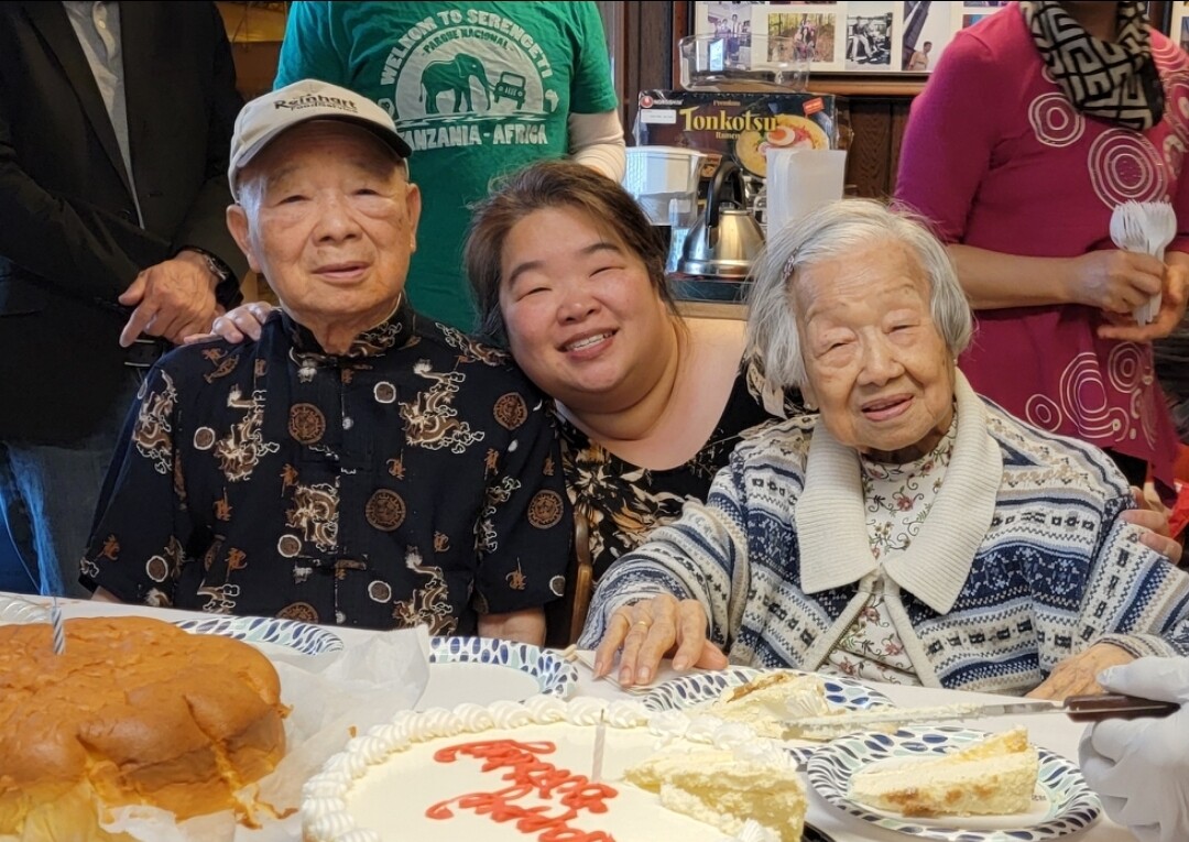 Three generations at a birthday table: two elderly grandparents with a smiling woman between them, cake and slices in foreground.