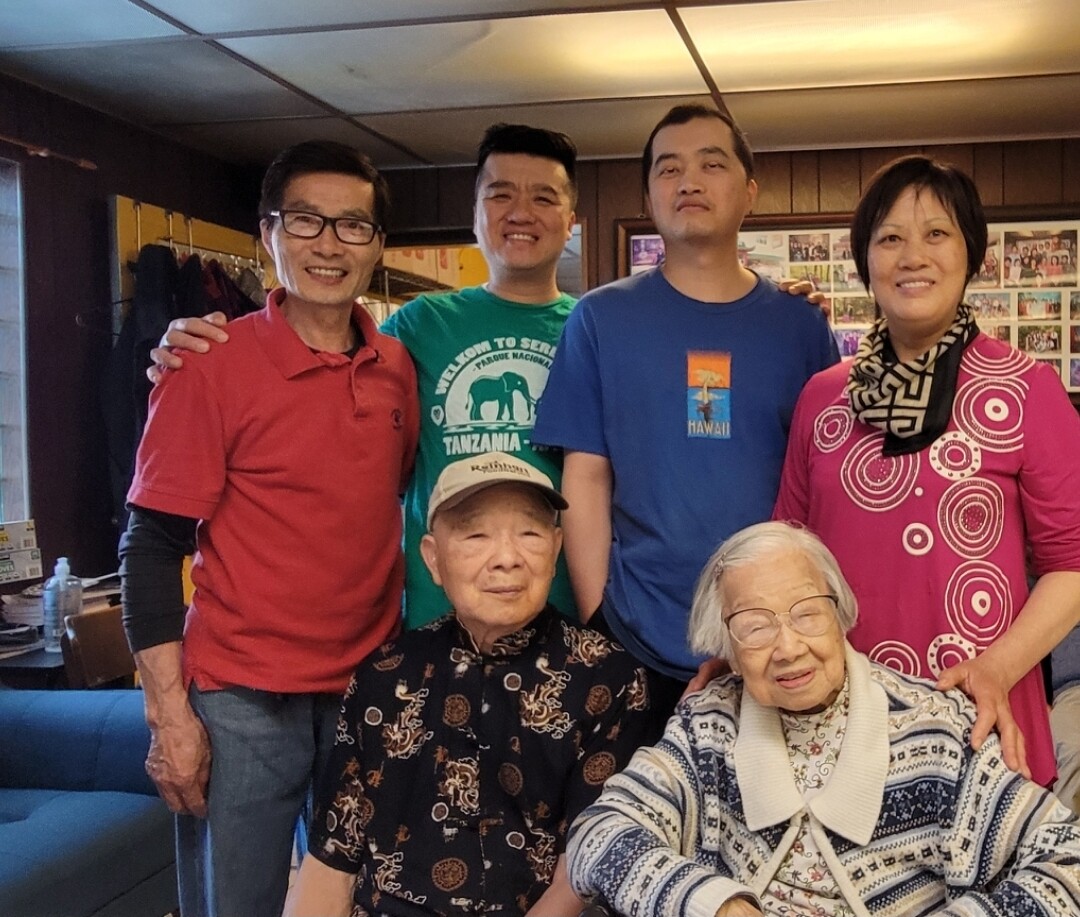 Six people pose together indoors: two elderly individuals sit at the front, smiling, while four adults stand behind them, also smiling; a cozy room with wood-paneled walls and a photo collage is in the background.
