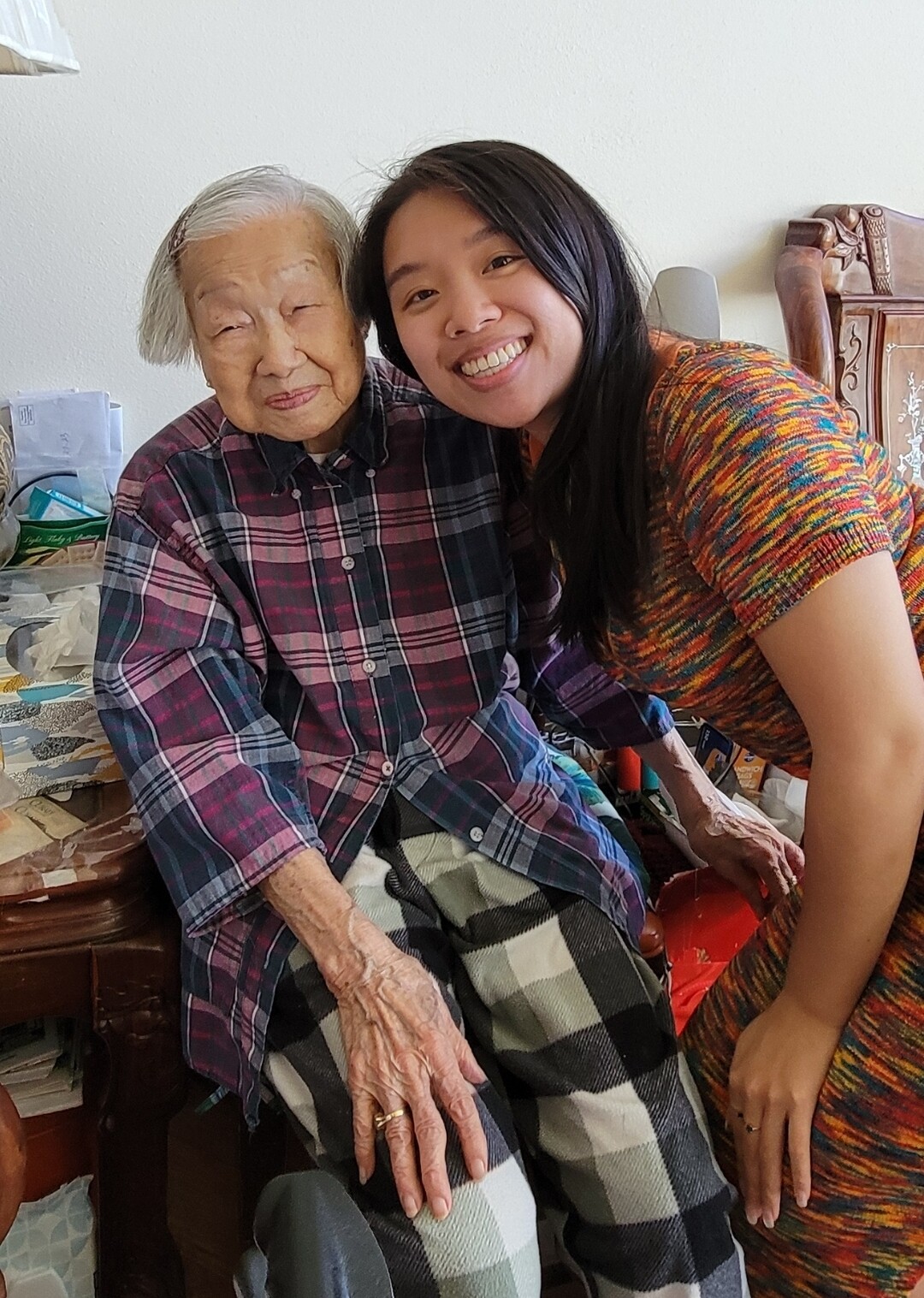 Young woman leaning in and smiling with her elderly grandmother who sits in a plaid shirt at home.