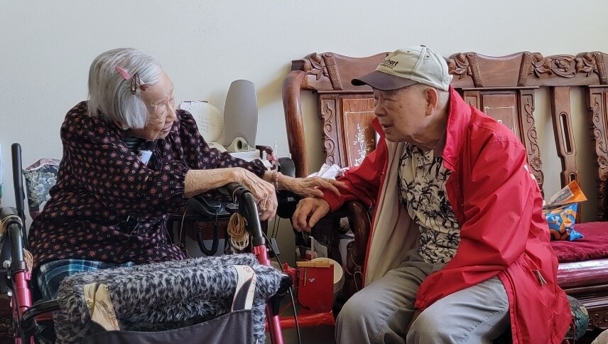 Two elderly people sit together indoors; a woman in a wheelchair holds hands with a man in a red jacket, sharing a conversation in a living room.