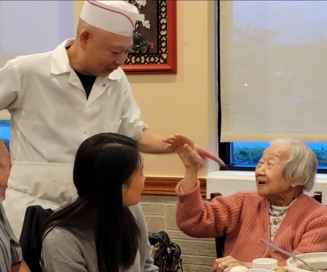Nurse or chef in white uniform offering a gesture to an elderly woman at a dining table, who smiles and raises her hand in response.