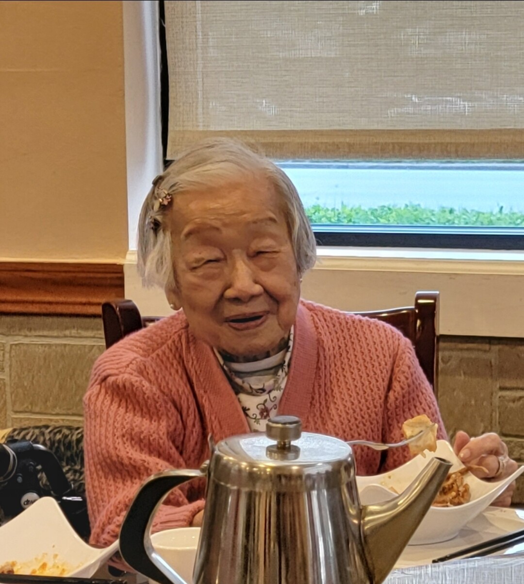Elderly woman with short white hair in a pink cardigan sits at a dining table with a shiny metal kettle in front of her.