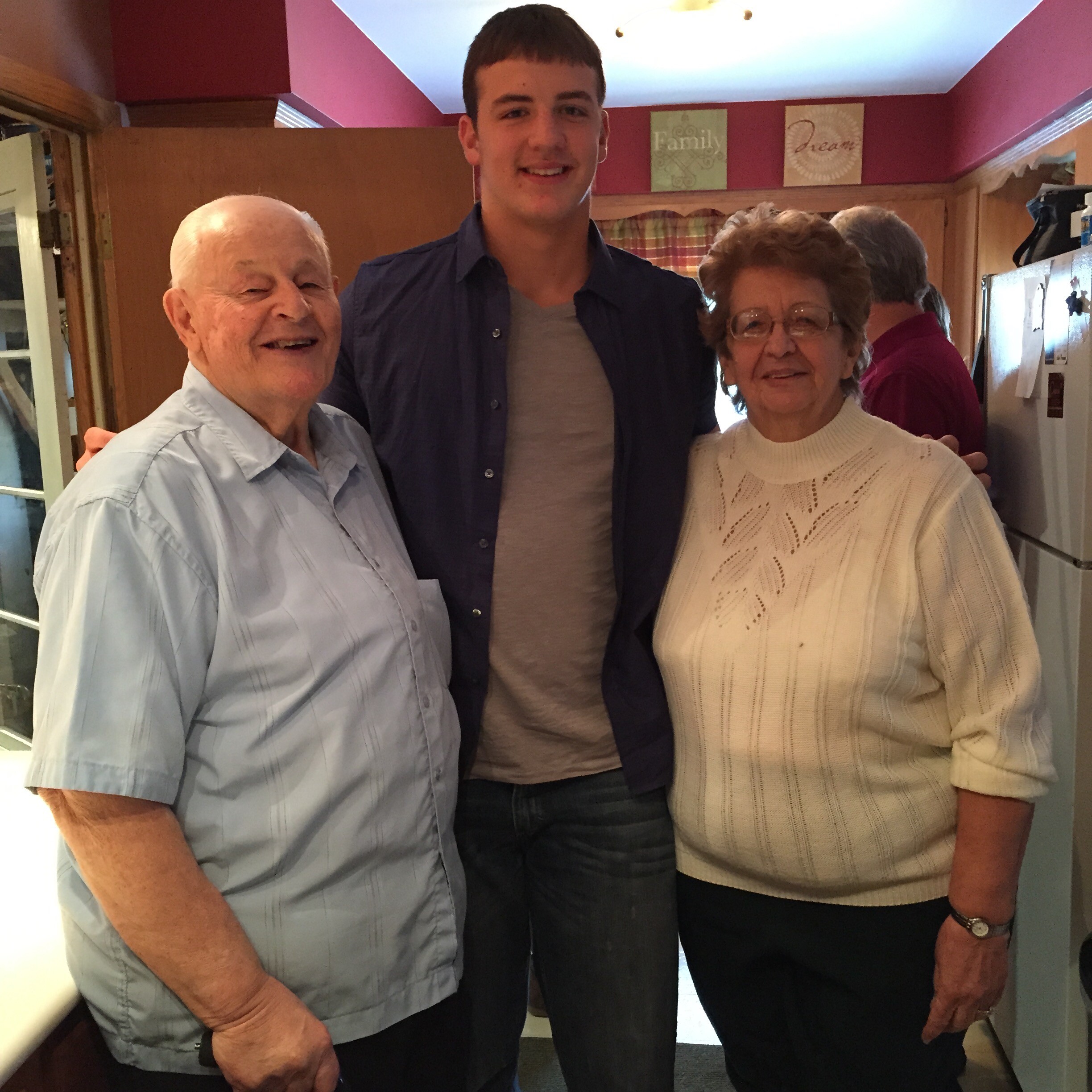 Three adults posing together indoors, smiling at the camera in a cozy home setting with warm red walls behind them in a kitchen area.