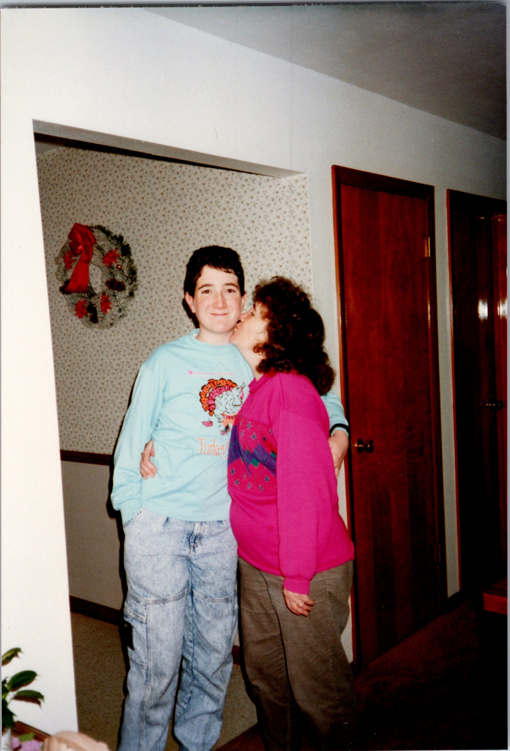 Two people share a hug indoors: a man in a light turquoise shirt and a woman in pink, with a holiday wreath on the wall behind them.