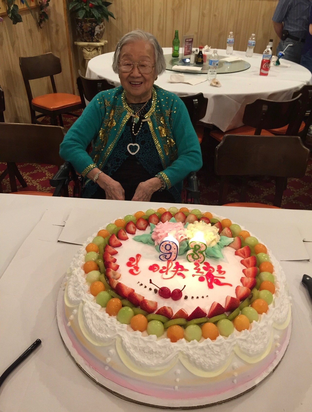 Smiling elderly woman in a turquoise embroidered jacket sits at a table with a large fruit-topped birthday cake in front of her.