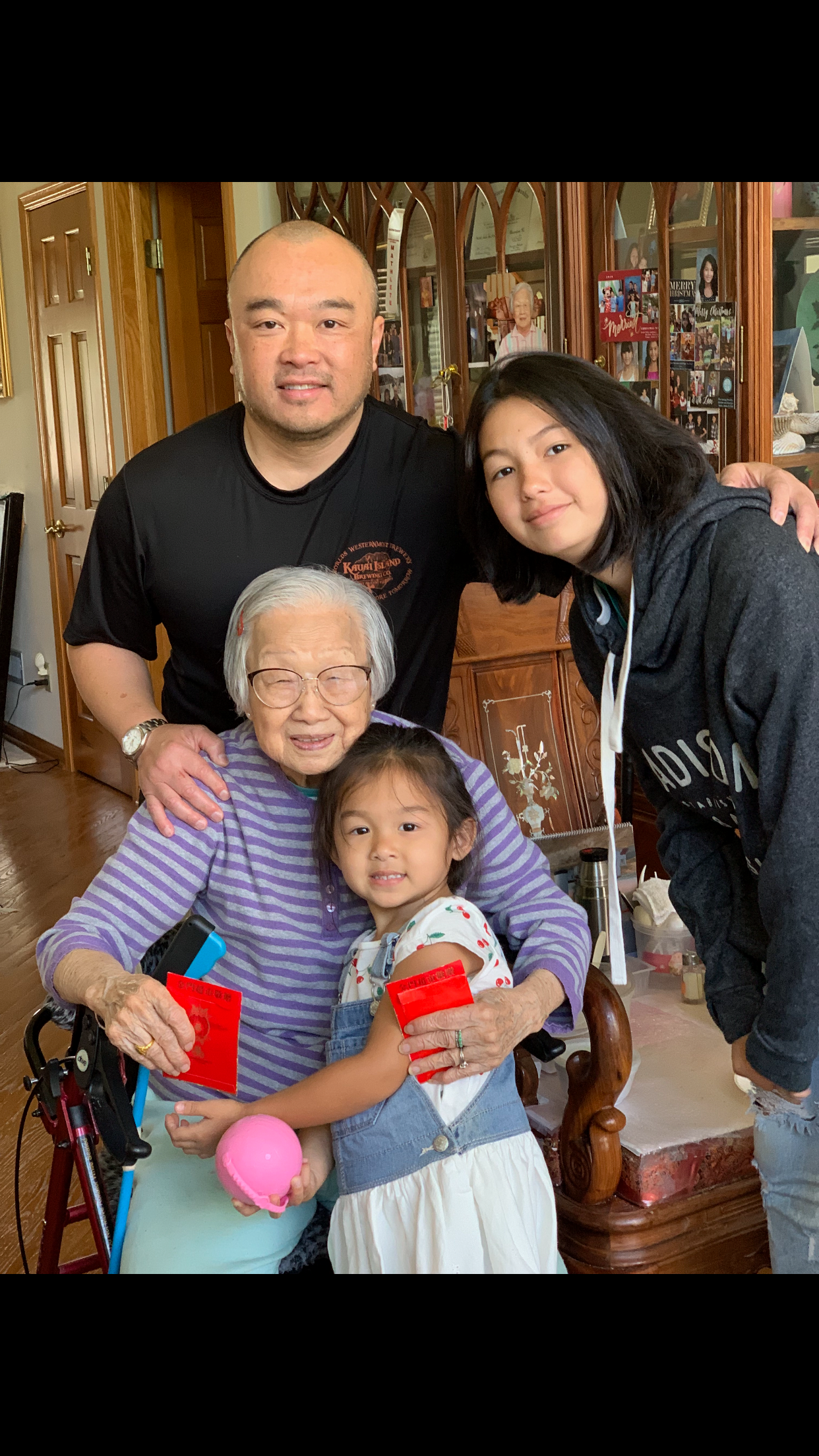 Grandmother in a wheelchair holding red envelopes, with a young girl in a denim dress and two adults smiling beside them indoors.