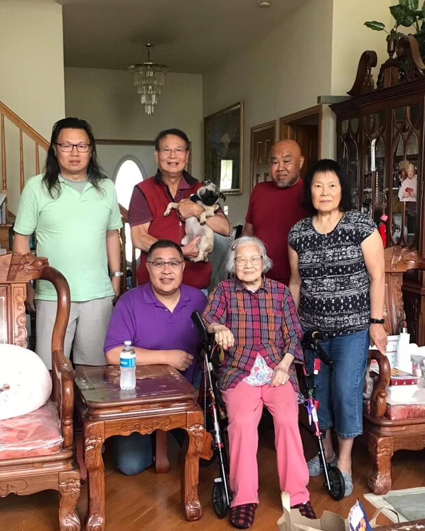 Multi-generational family portrait in a living room; an elderly woman in a wheelchair sits front center, with relatives and a dog nearby.