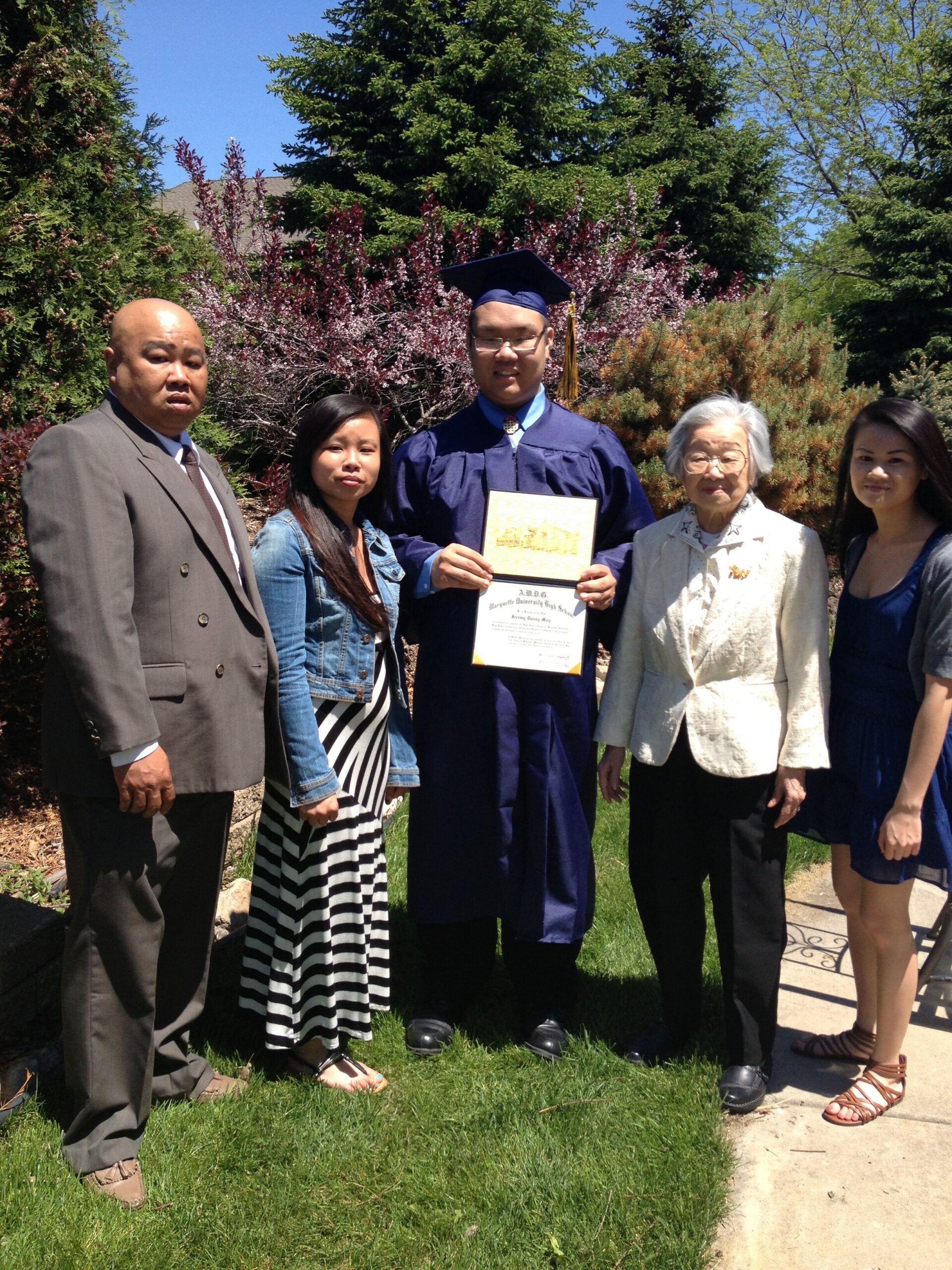 Graduate in navy cap and gown holding diploma outdoors, flanked by family members in spring yard.