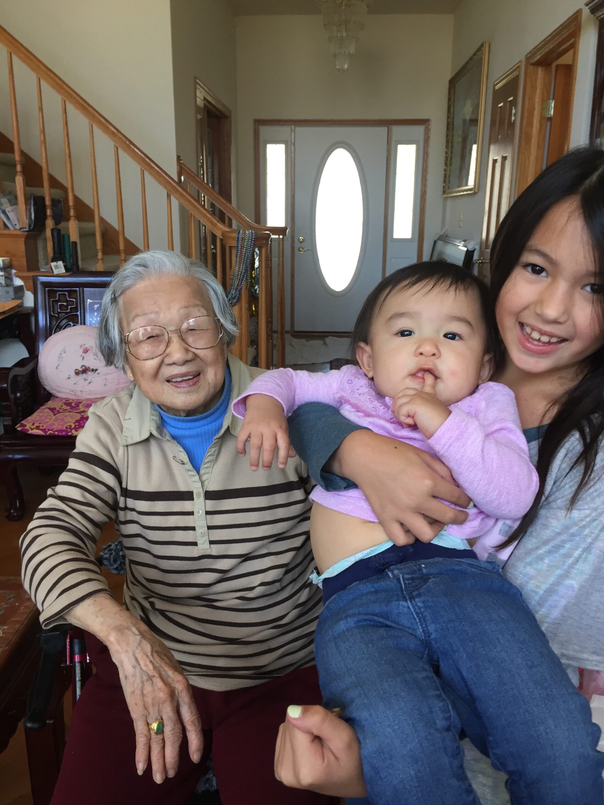 Smiling elderly woman with glasses sits beside a young girl holding a baby in a home living room. Stairs and a front door are in the background.