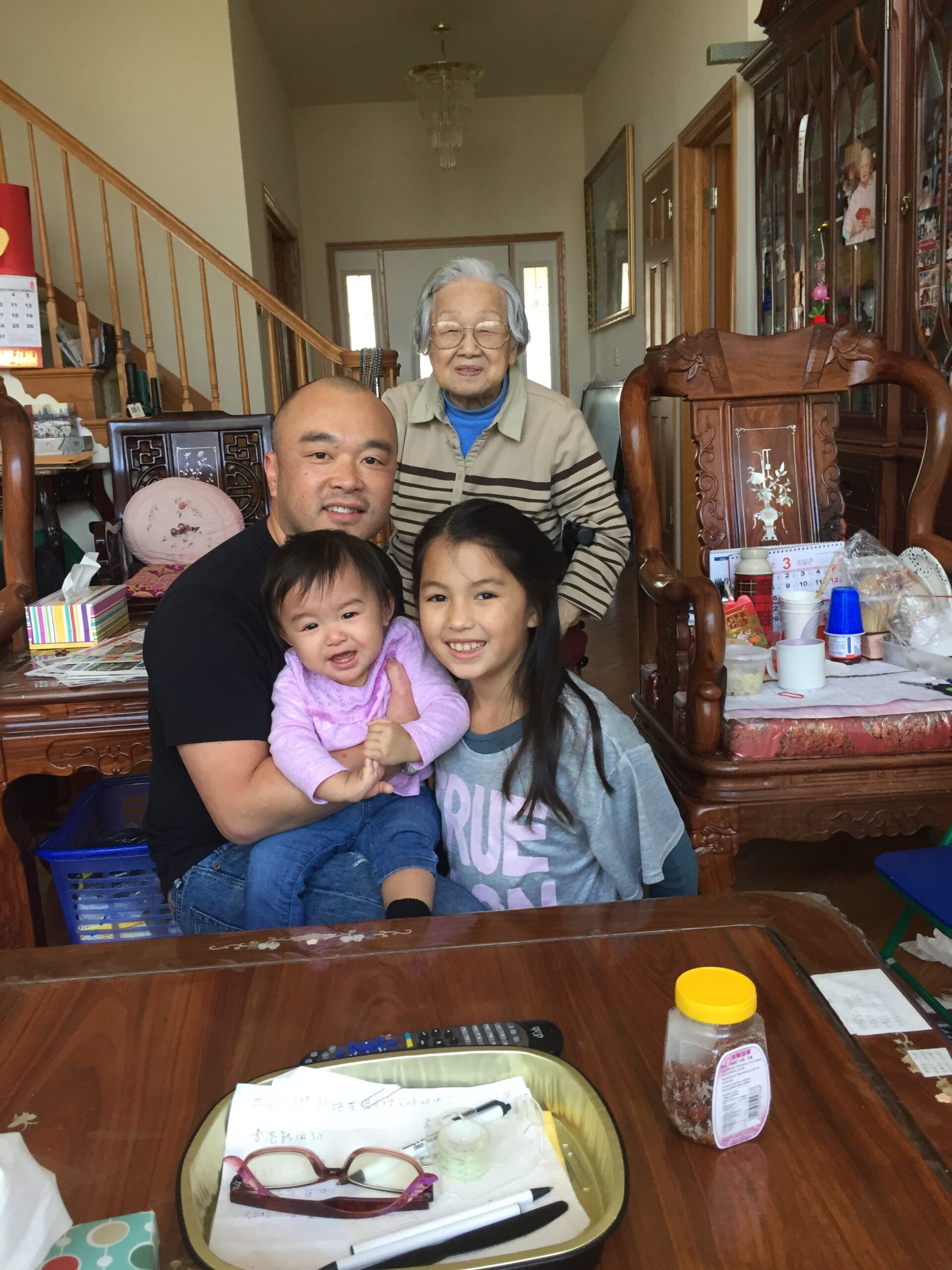 A man sits with two young children on his lap as an elderly woman stands behind them in a living room filled with wooden furniture and family photos on display.