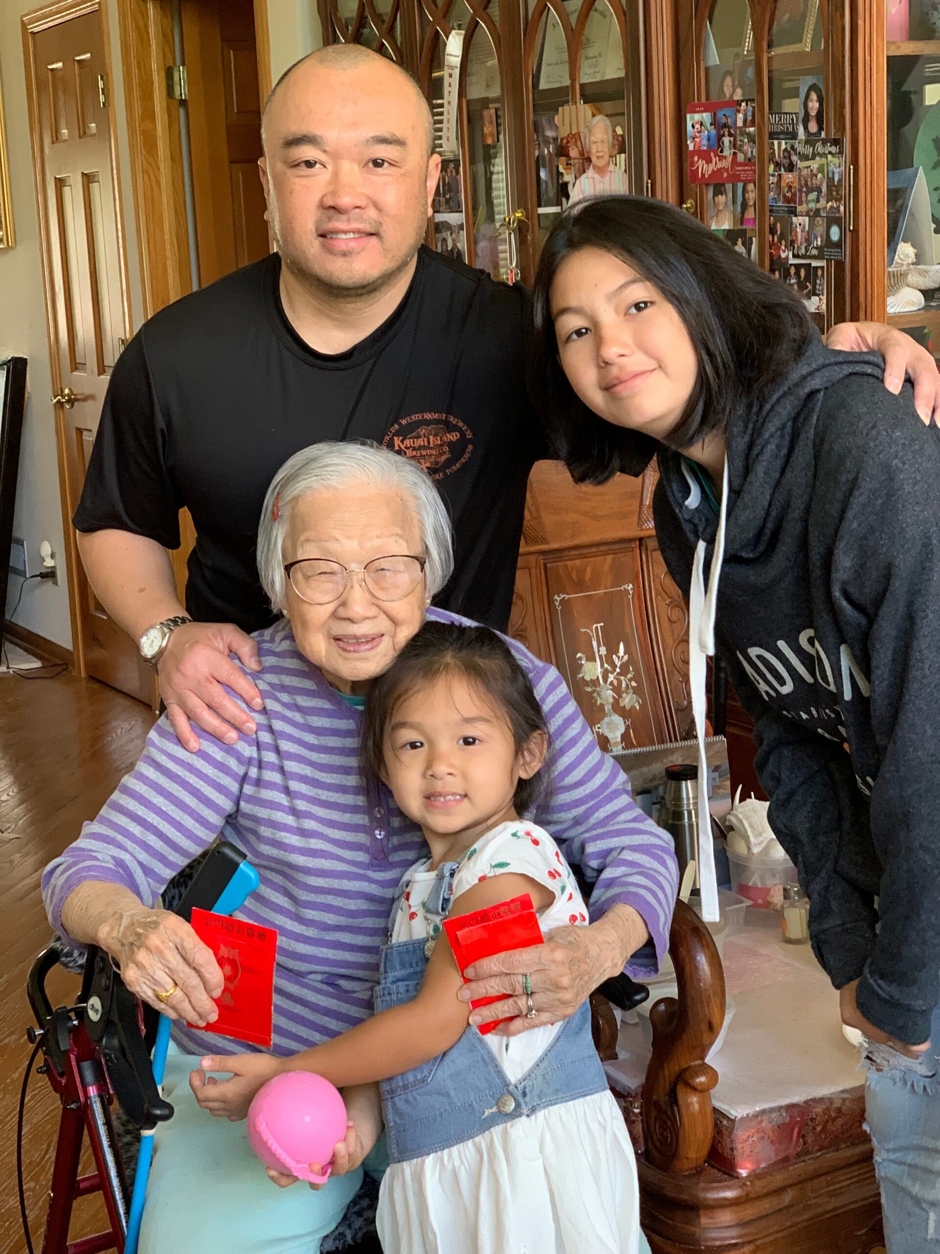 Elderly woman in a wheelchair next to a young girl, both holding red envelopes as a man and another young woman lean in and smile in a home setting.