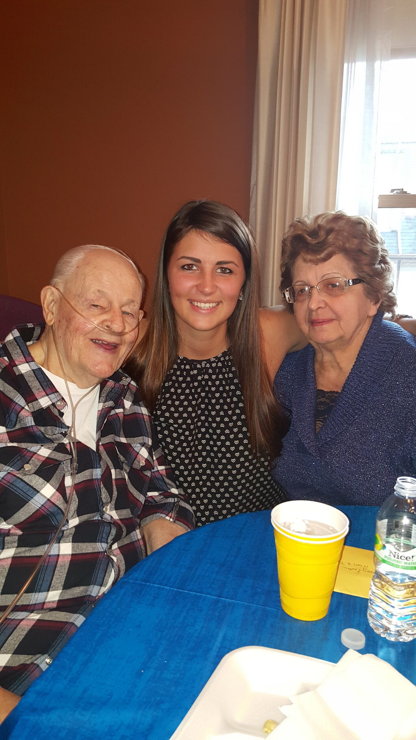 Three people sit together smiling: an elderly man with a nasal oxygen tube, a young woman in the middle, and an older woman on the right at a blue table with cups and a water bottle nearby