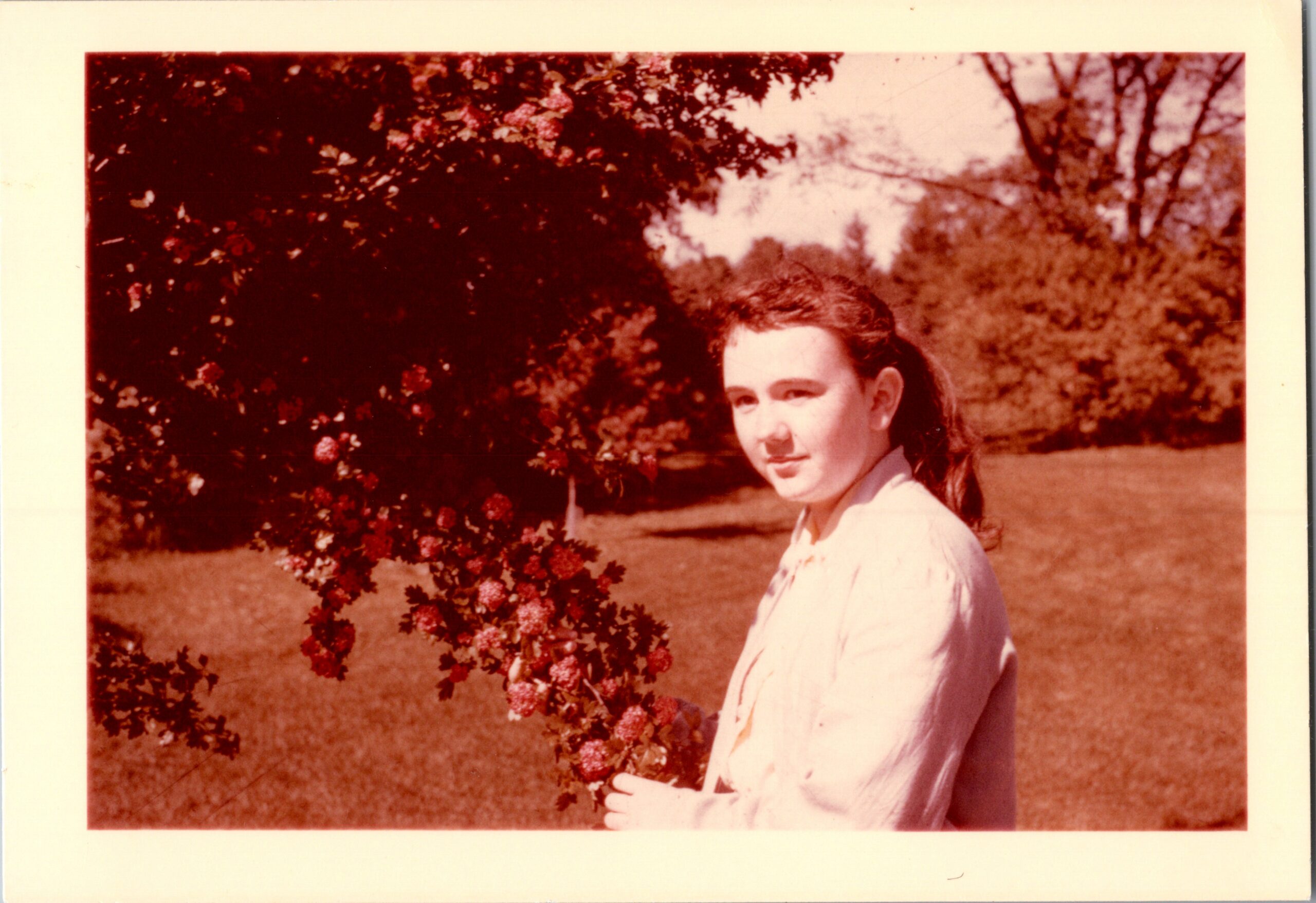 Young woman with dark hair in a light blouse stands beside a flowering bush in a sunny park setting, facing slightly toward the camera.