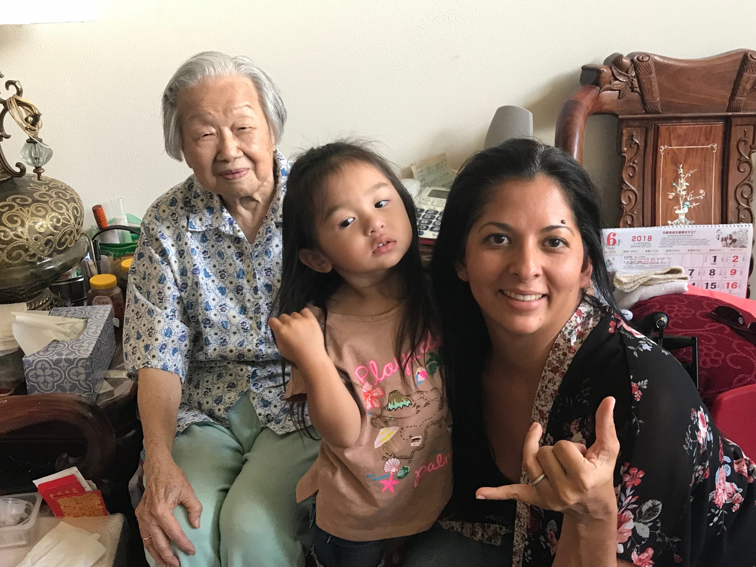 Three generations posing for a selfie indoors: an elderly woman in a blue floral shirt, a young girl, and a smiling woman in a floral top next to her.