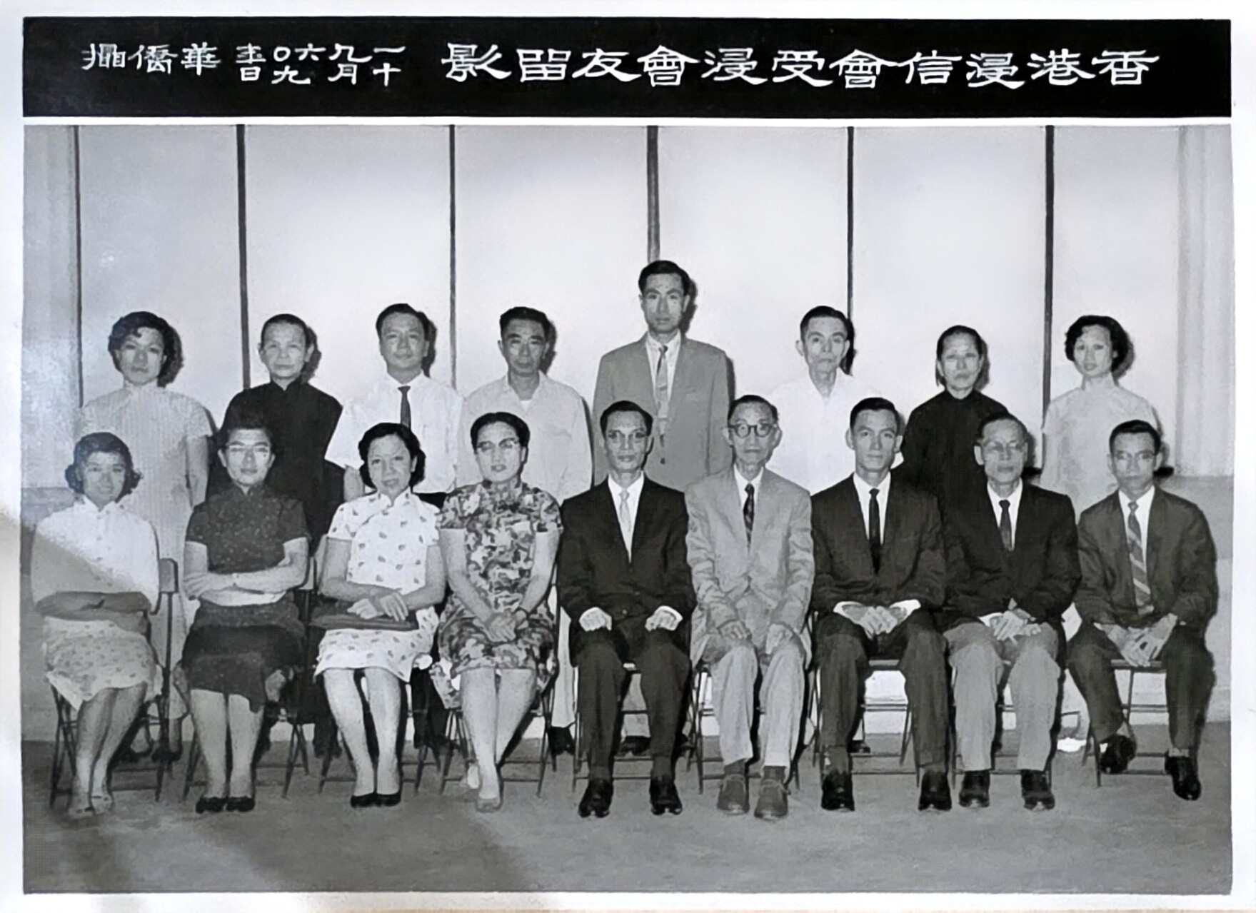 Group portrait of men and women in formal attire posing for a class or group photo, with a Chinese banner above and people arranged in two rows (seated front row, standing back row).