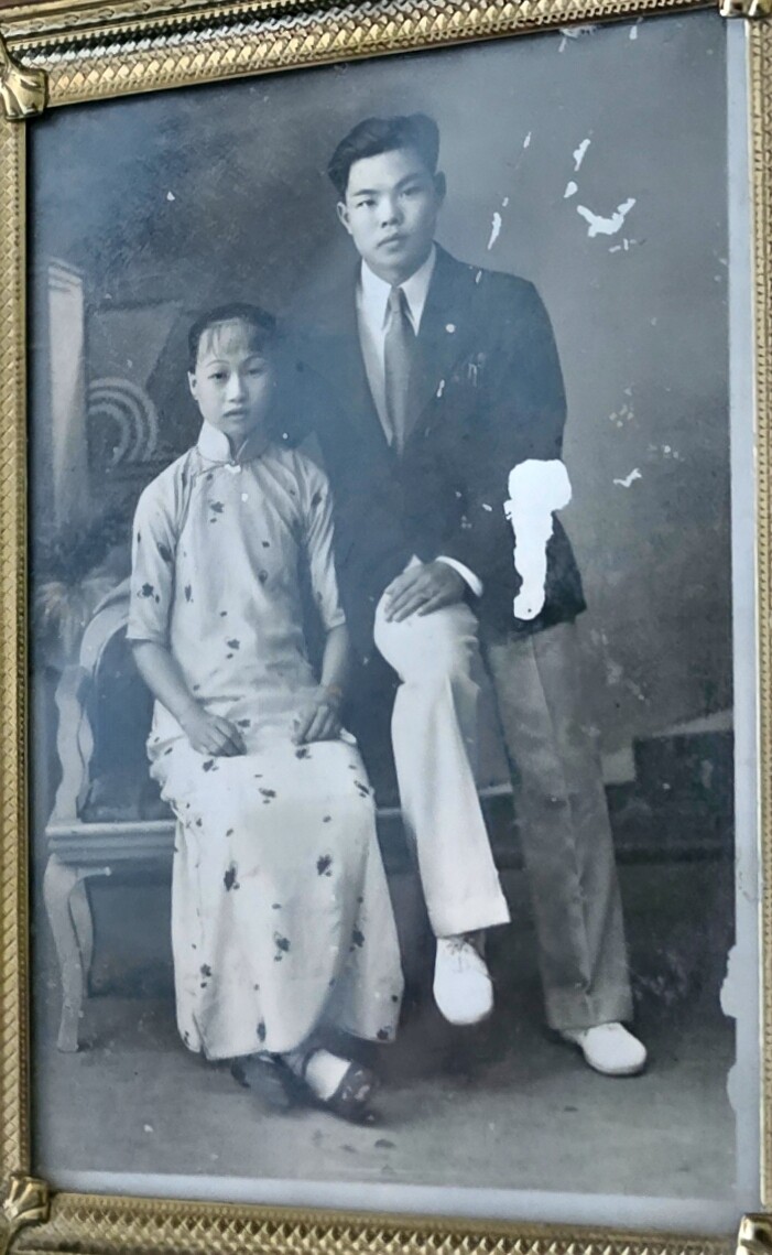 Studio portrait of a man in a suit standing beside a seated woman in a traditional cheongsam (qipao). They pose formally in a vintage photograph, both looking at the camera.