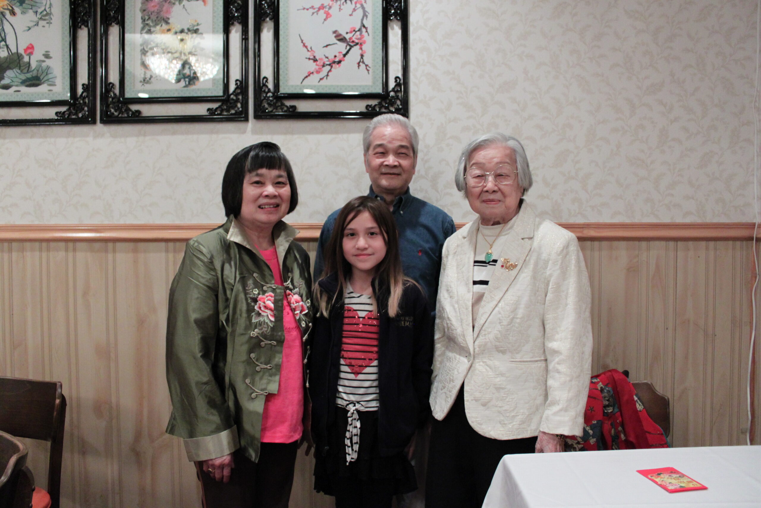 Group portrait of four people—a young girl, a middle-aged man, and two elderly women—standing together indoors with framed art on the wall behind them and a patterned wallpaper backdrop.