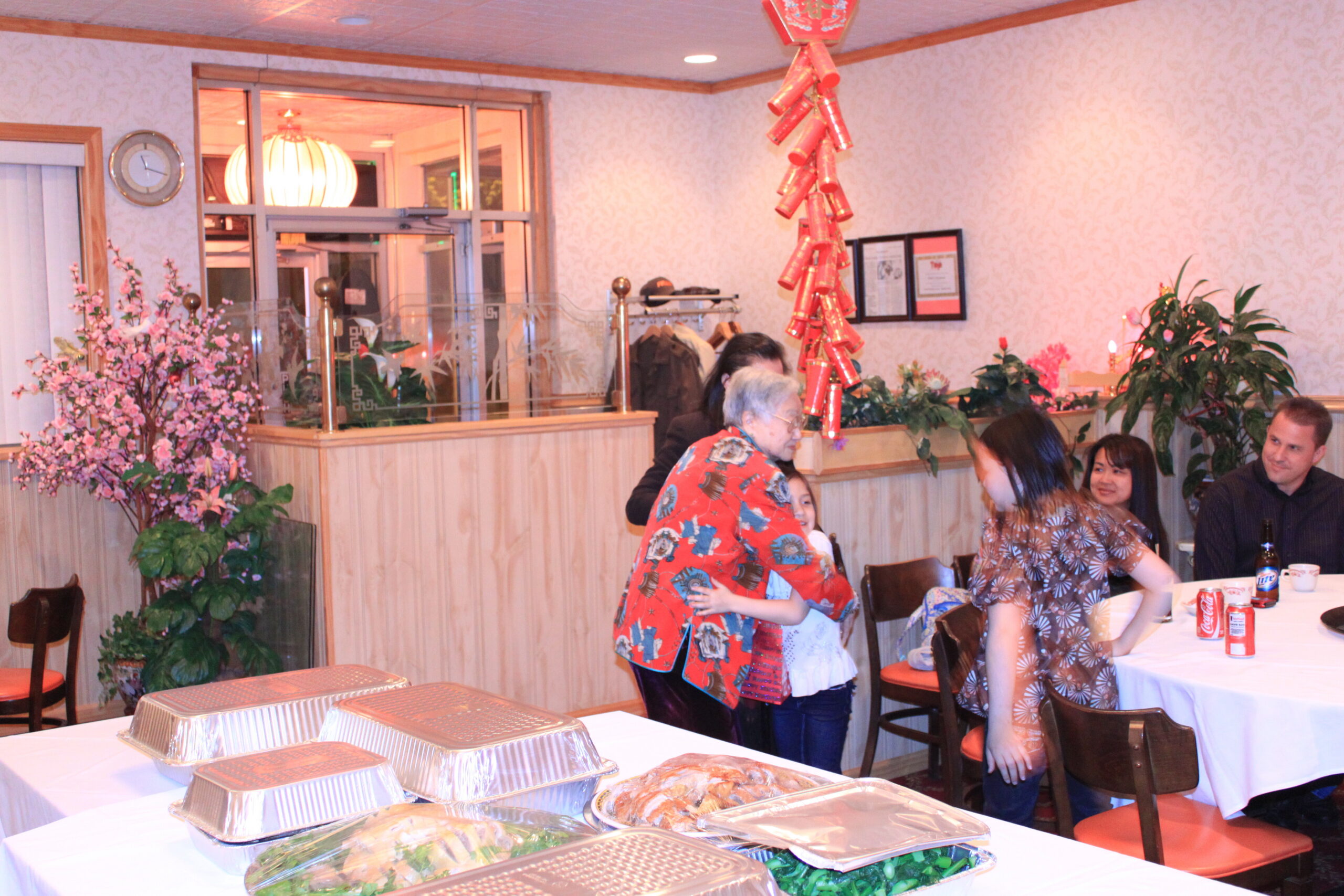 People at a festive restaurant party with tables full of aluminum catering trays and red decorations hanging from the ceiling.