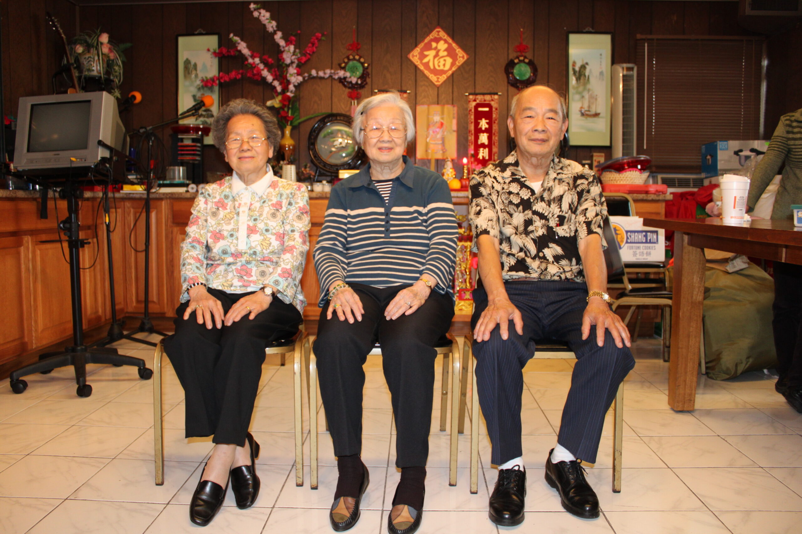 Four older adults seated in a row on chairs, smiling, in a festively decorated room with wood paneling and decorations behind them.