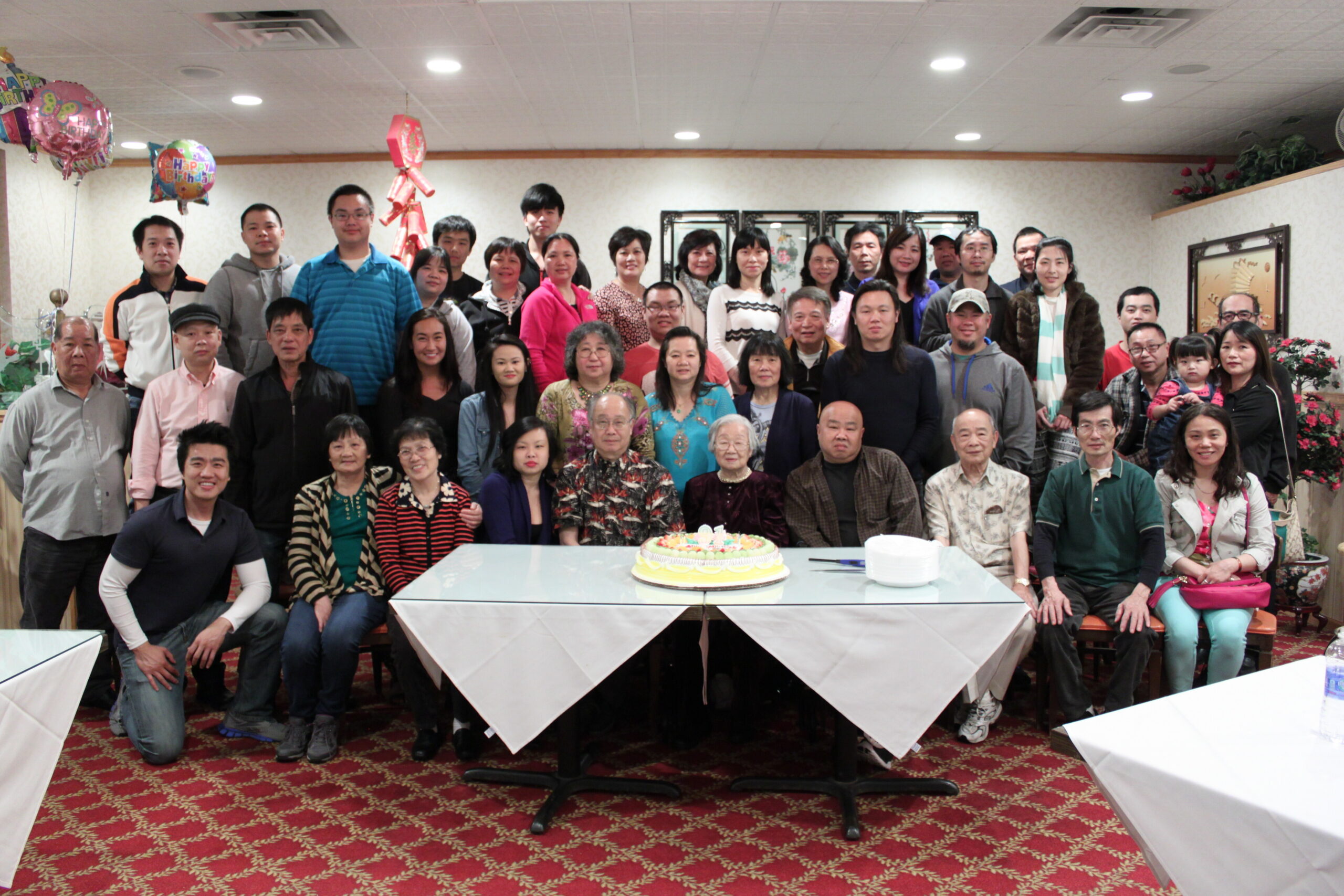 Large group of people gathered in a birthday party room around a table with a cake in the foreground, balloons visible.