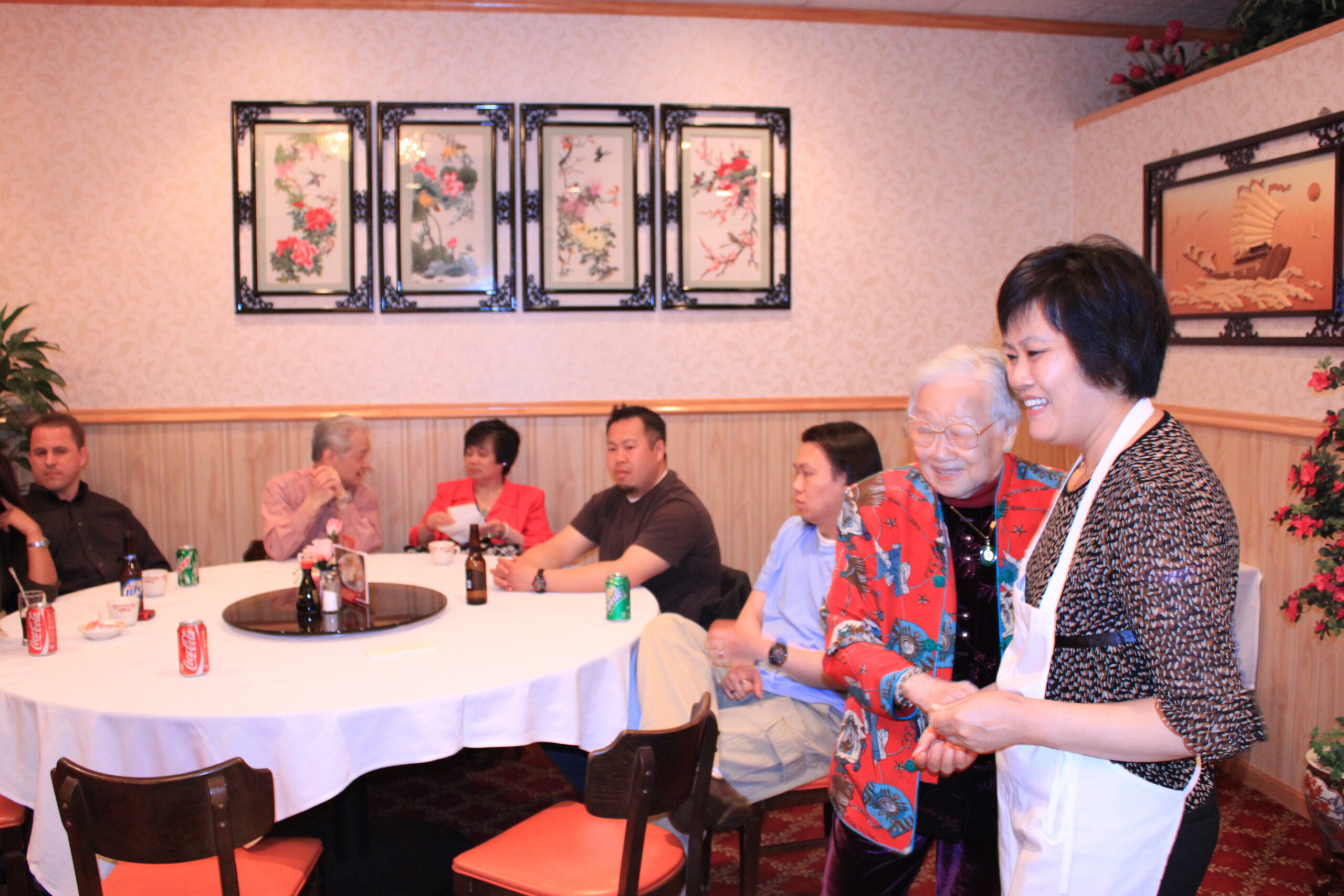 Several guests sit around a round banquet table with drinks while two women in foreground smile; one wears a colorful scarf and apron at a social gathering in a restaurant interior with framed art on the wall.