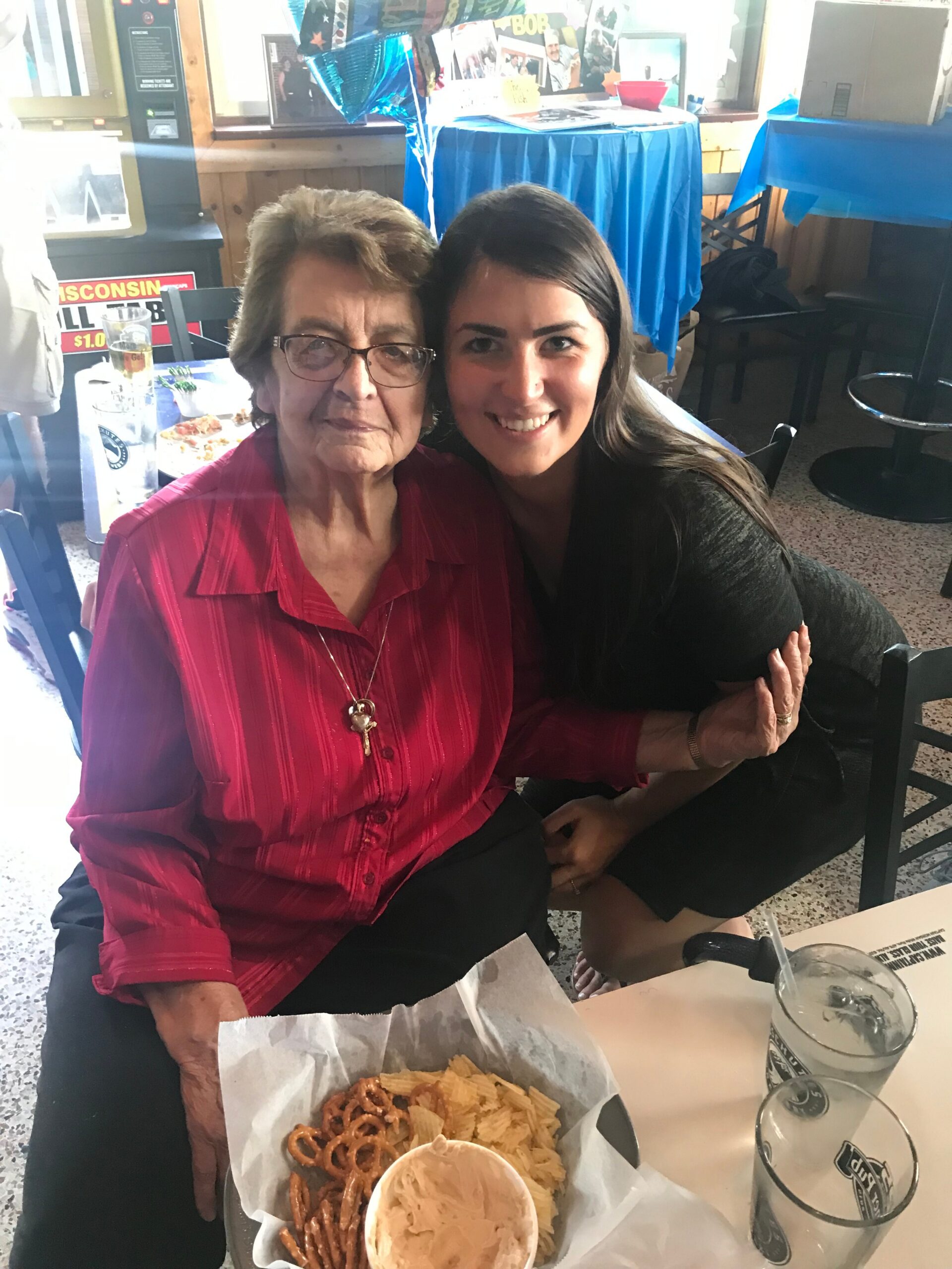 Two women hug at a table in a casual restaurant; elderly woman in red shirt beside younger woman smiling, with snack tray and drinks in front.