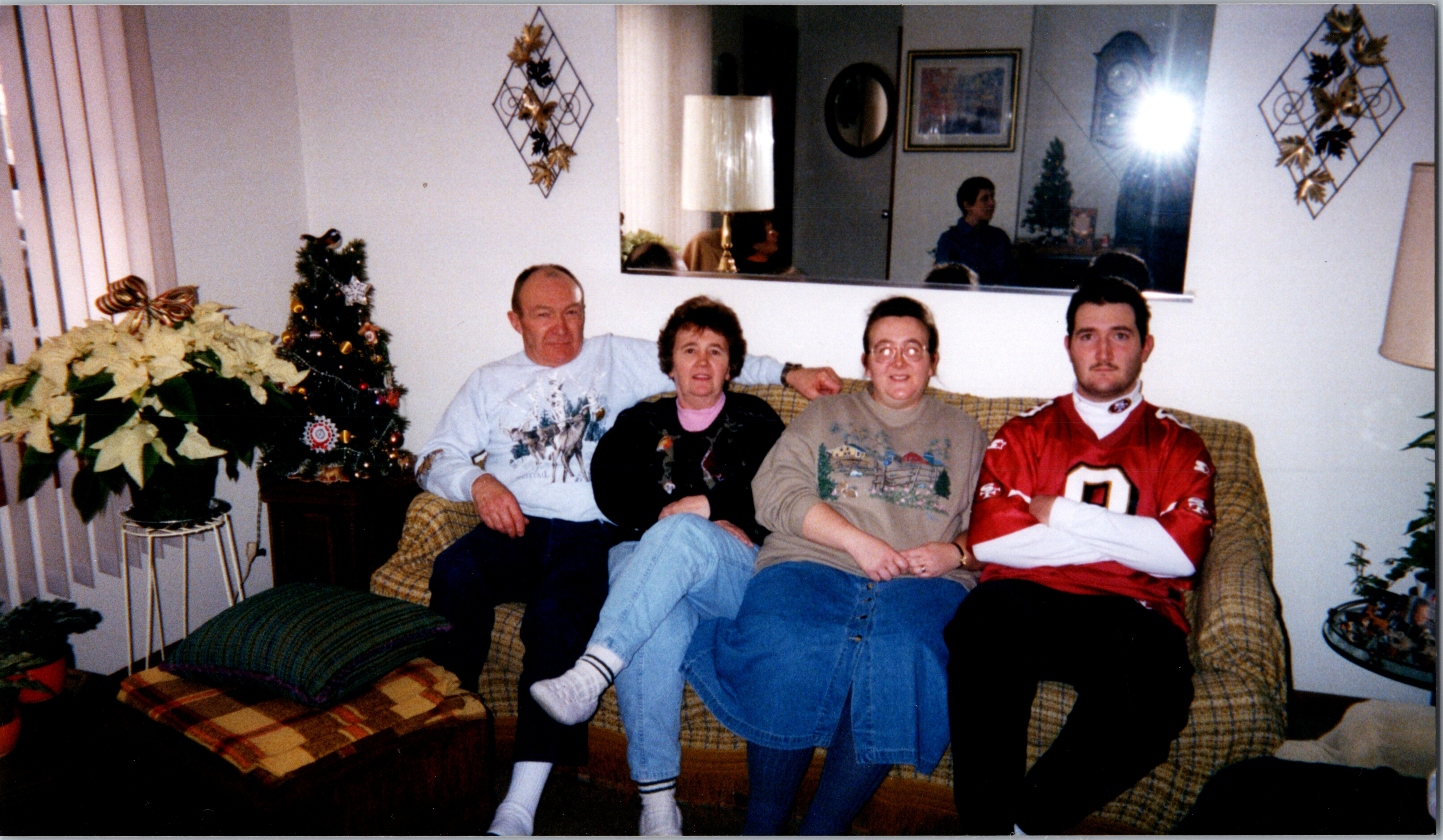 Four people sit together on a plaid sofa in a cozy living room decorated for Christmas, with a small tree and poinsettias nearby.