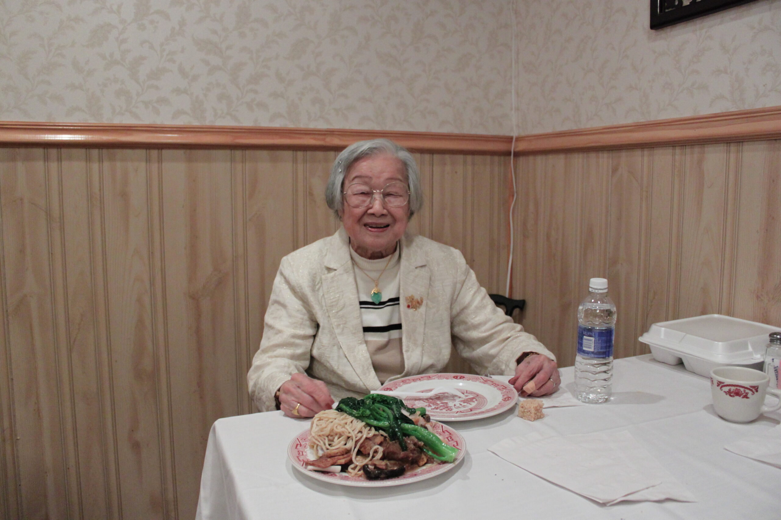 Older woman with glasses seated at a table, smiling, with plates of noodles and greens in front of her, plus a water bottle and takeout containers on a white tablecloth.
