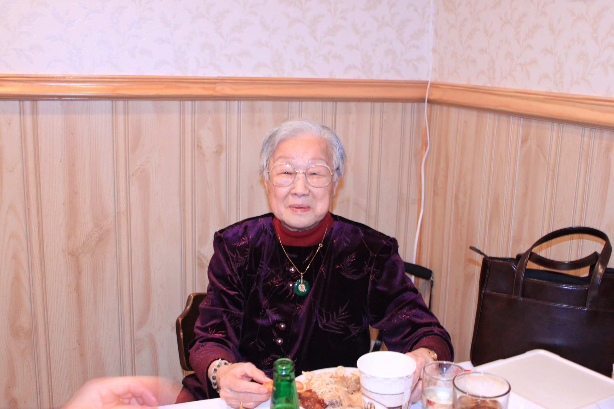 Older woman with short gray hair and glasses sits at a dining table, wearing a dark purple top with a green pendant, plates and cups in front of her on a wood-paneled wall backdrop.