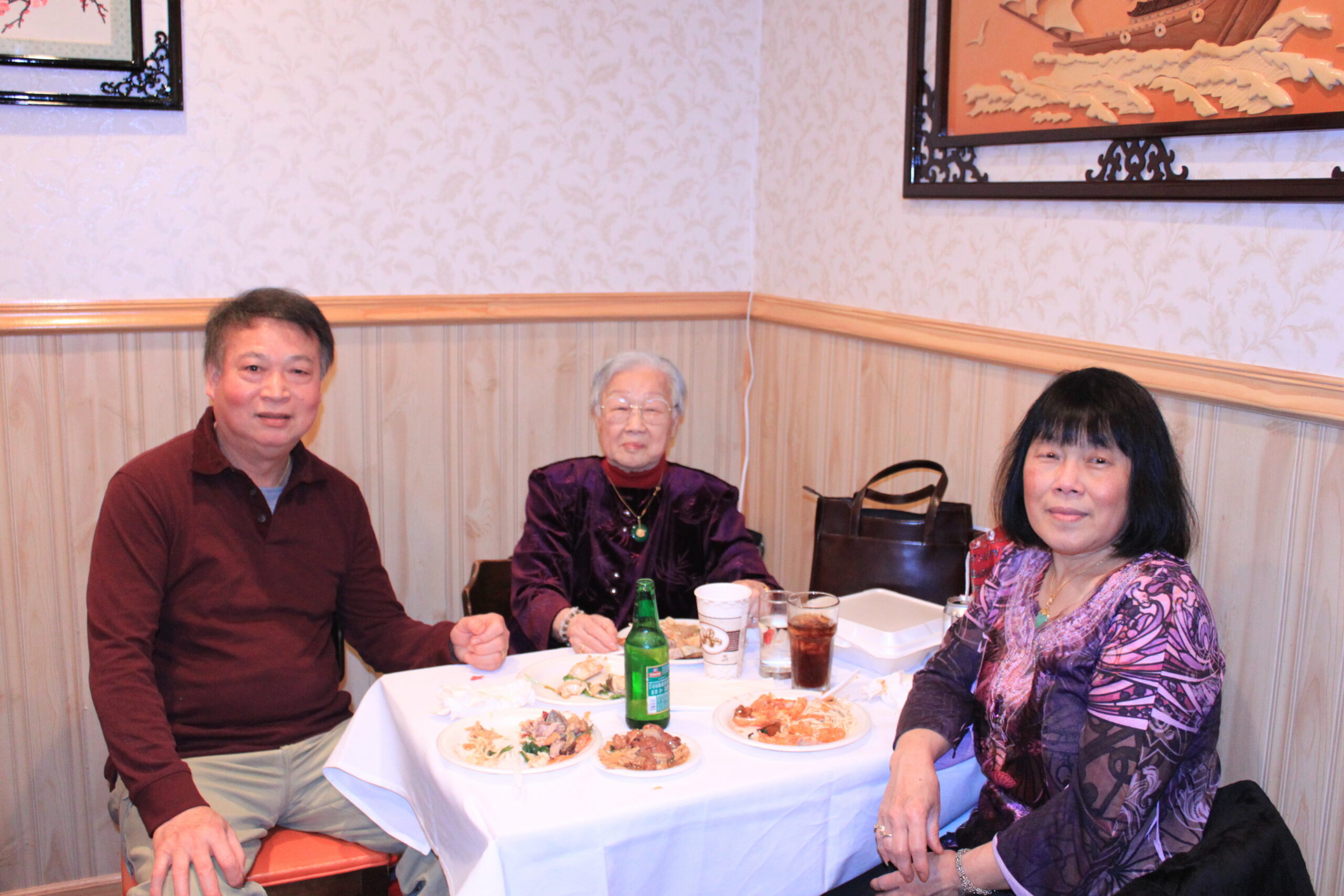 Three people sit at a dining table with plates of food and drinks in a casual restaurant setting.