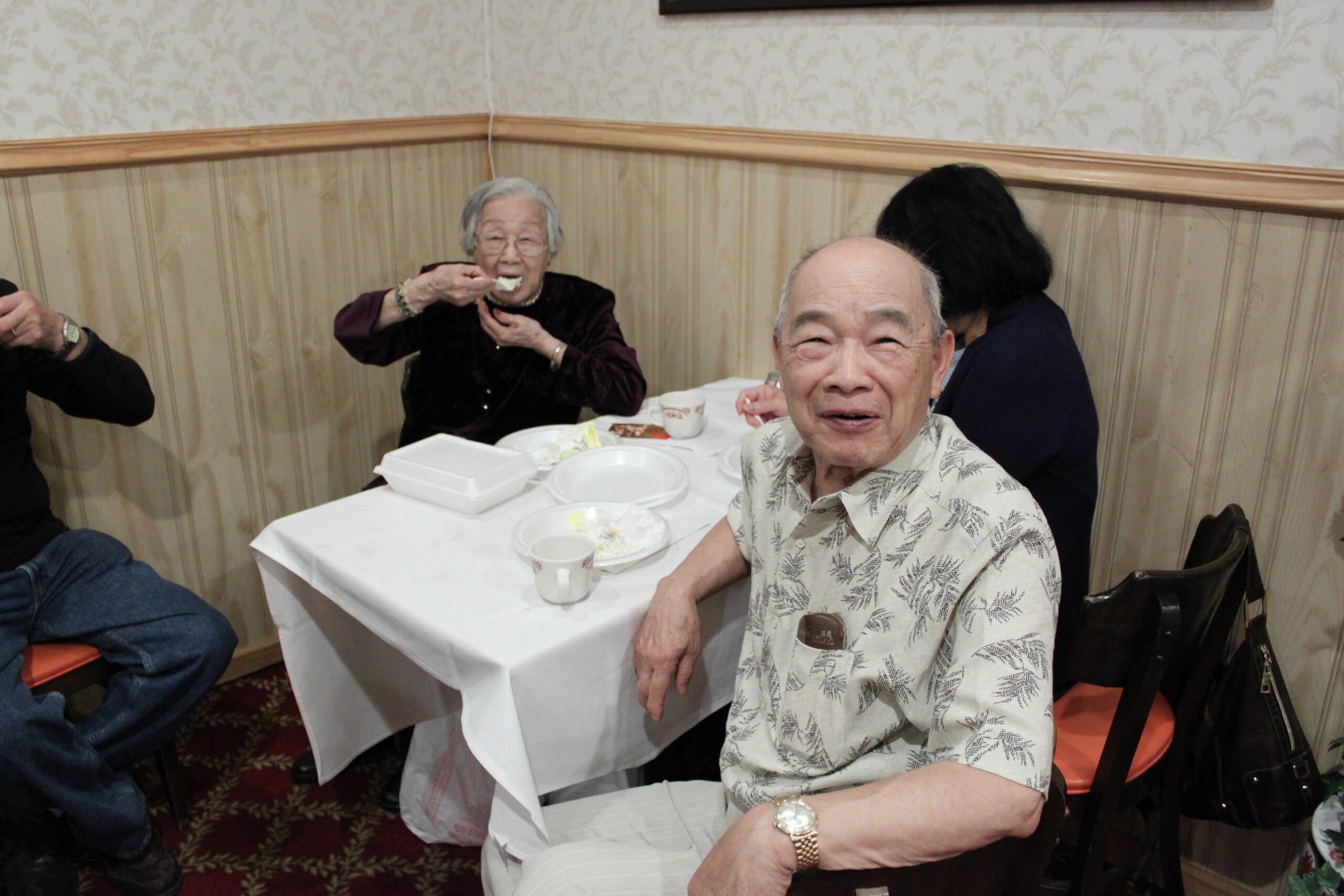Elderly man in a patterned shirt smiles at the camera while sitting at a dining table; an older woman in the background eats.