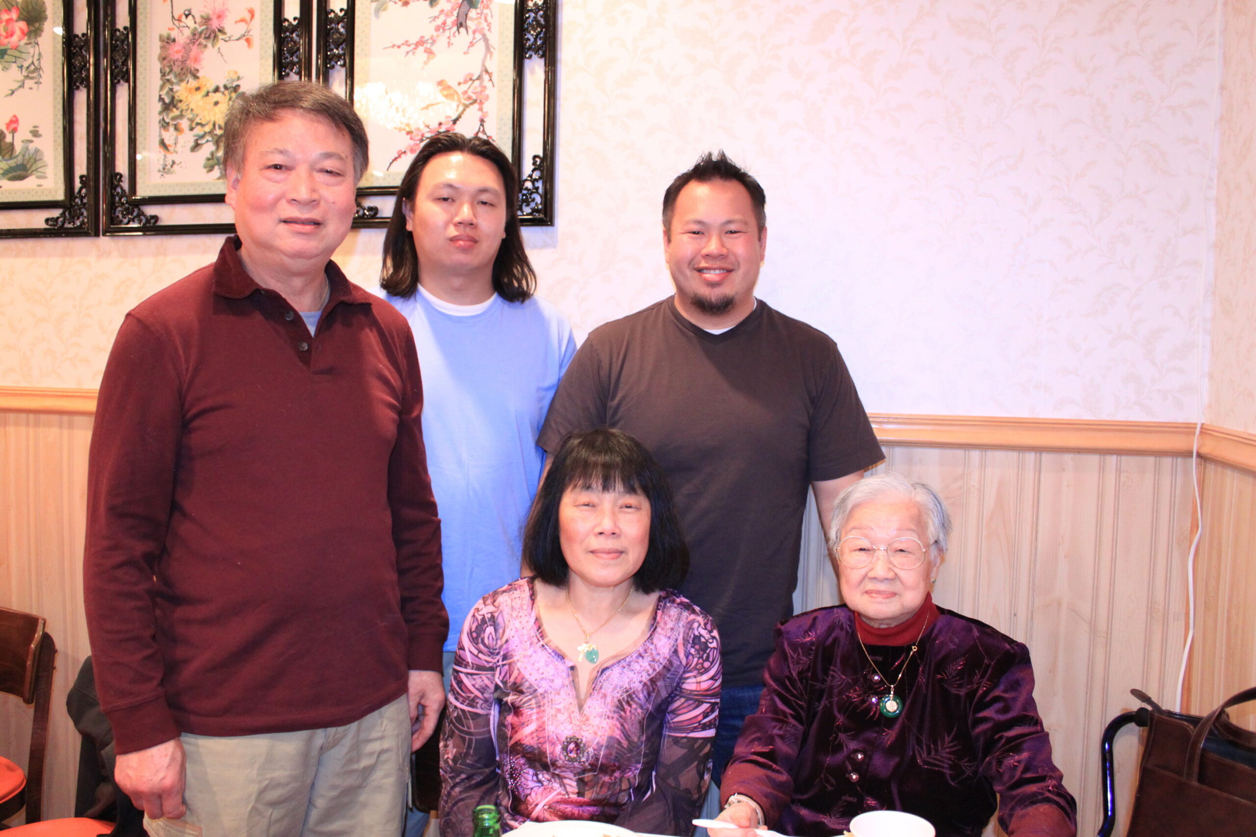 Six people, three standing and three seated, posing for a group photo in a restaurant with patterned wallpaper.