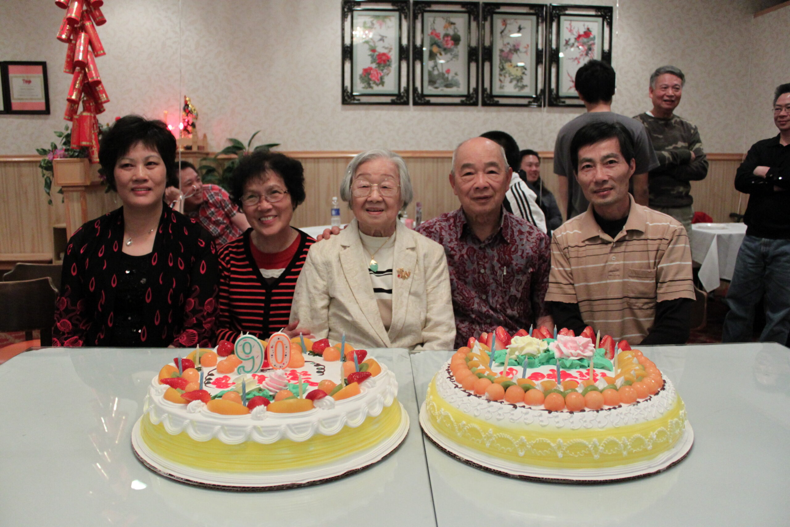 Group of adults at a birthday party with two fruit-topped cakes and colorful candles on a glass table in the foreground.