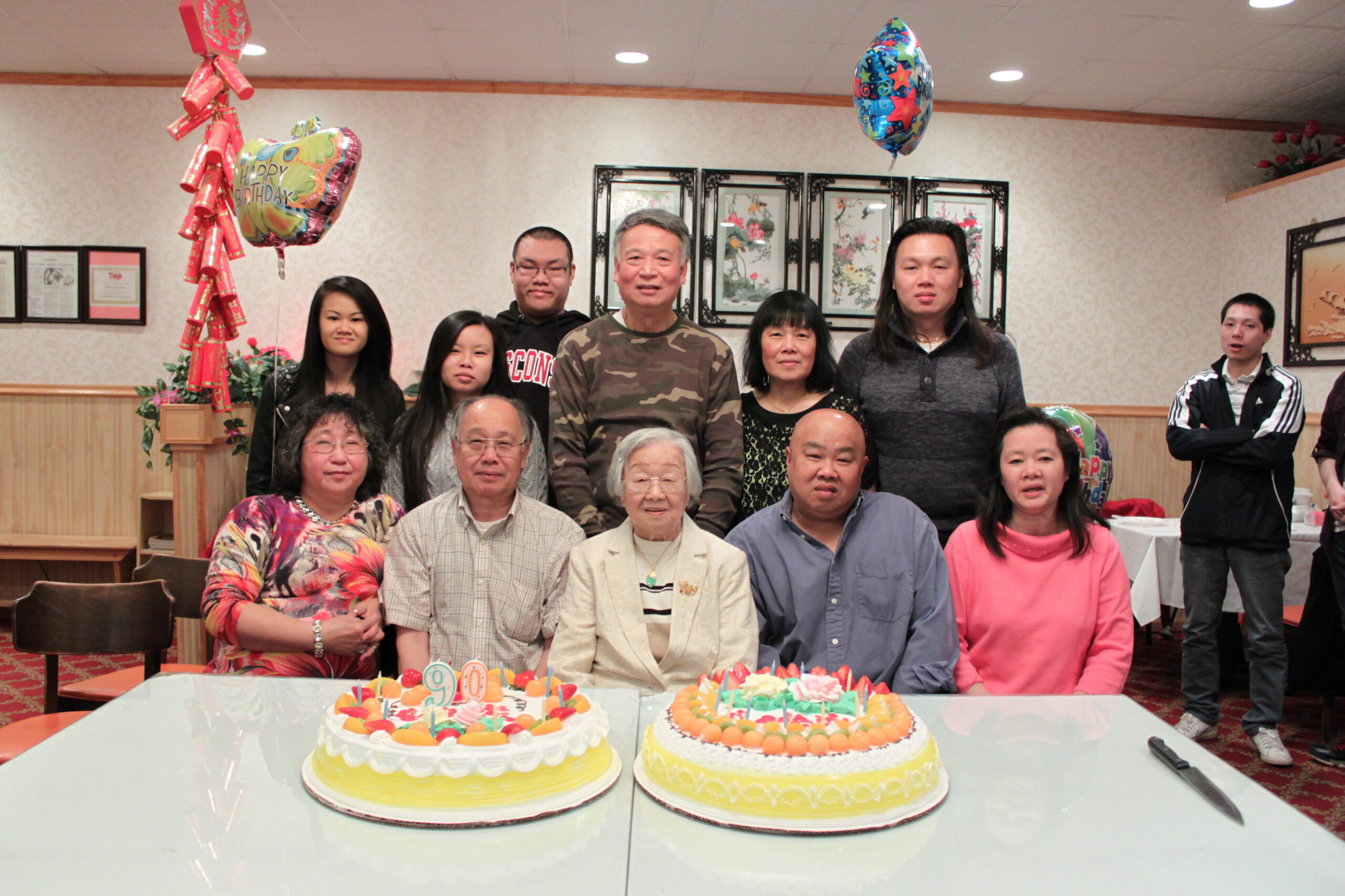 Group of smiling people posing around birthday cakes on a party table with balloons in a banquet hall.