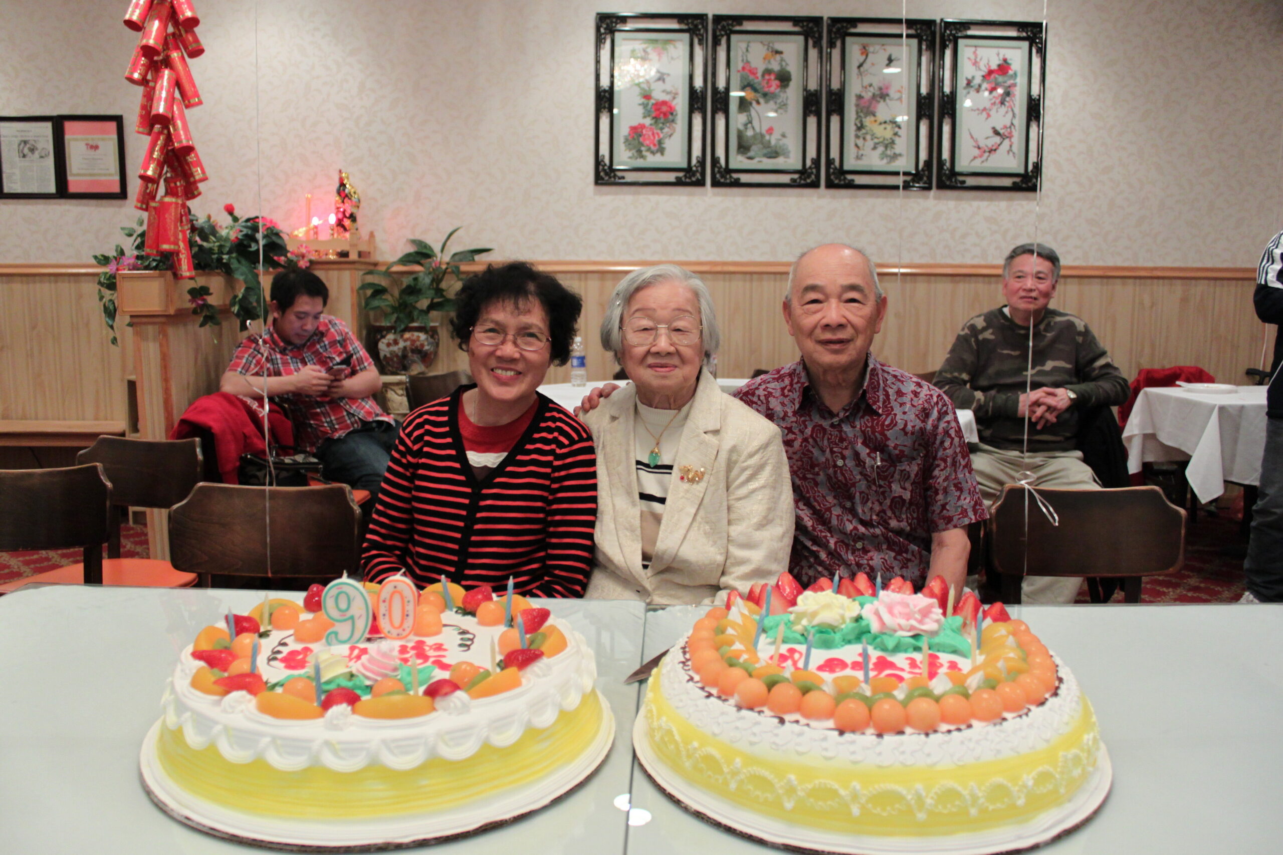 Four seniors smiling at a birthday celebration with two decorated cakes in the foreground and party decor in a dining hall.