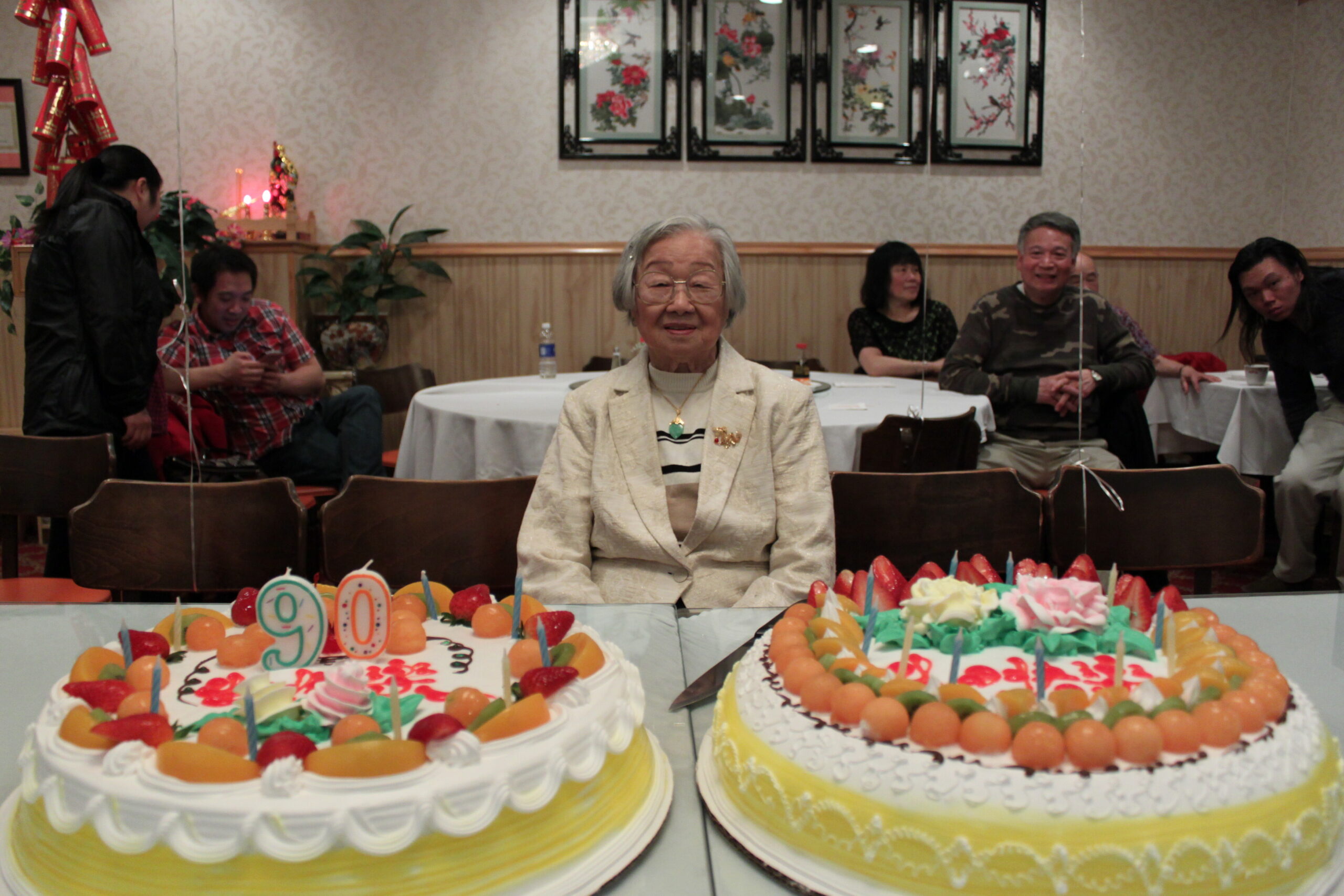 Smiling elderly woman in a light blazer sits at a table with two large fruit-topped birthday cakes in front.