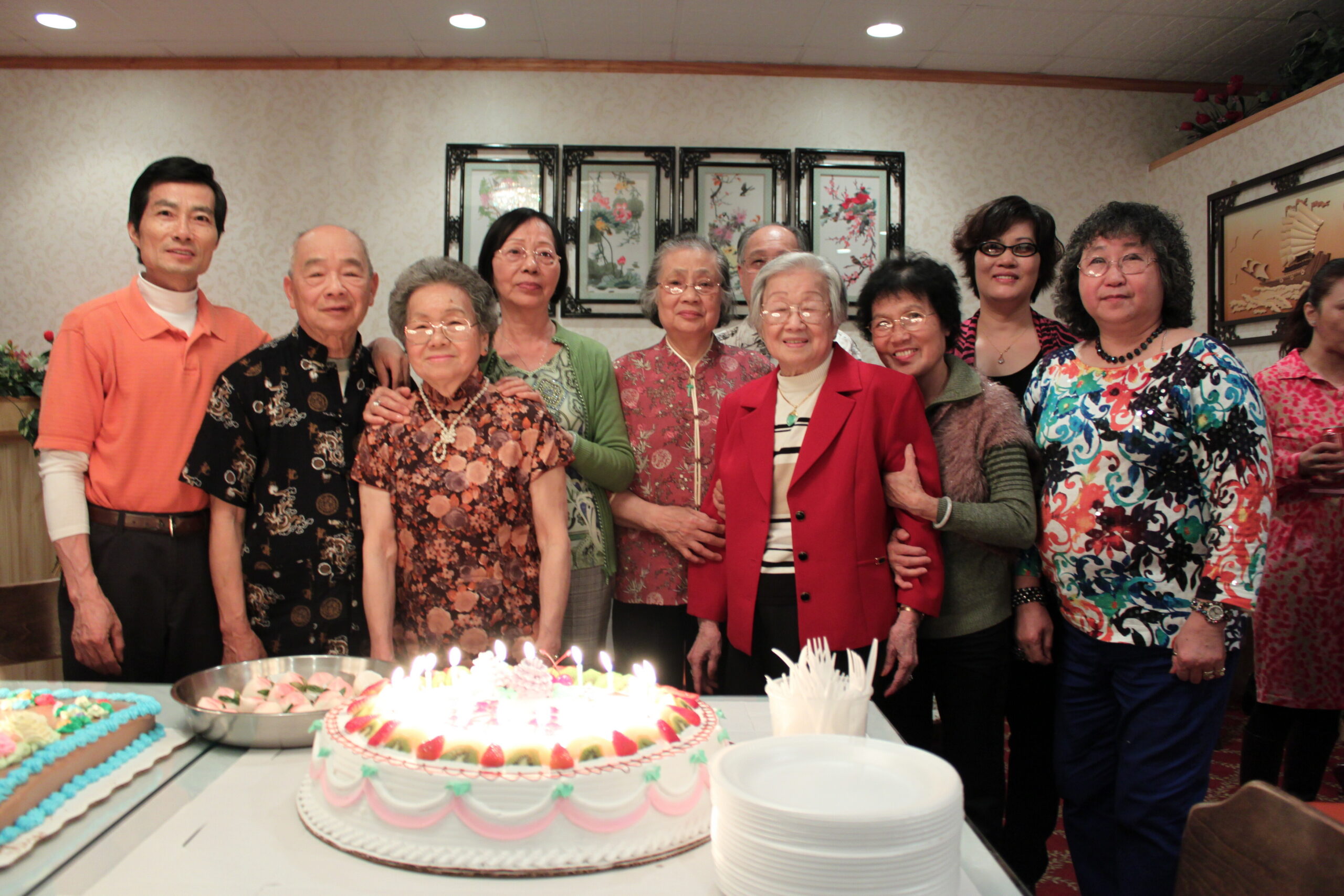 Group of diverse adults posing for a birthday party photo, standing behind a table with a large frosted cake and candles in front.