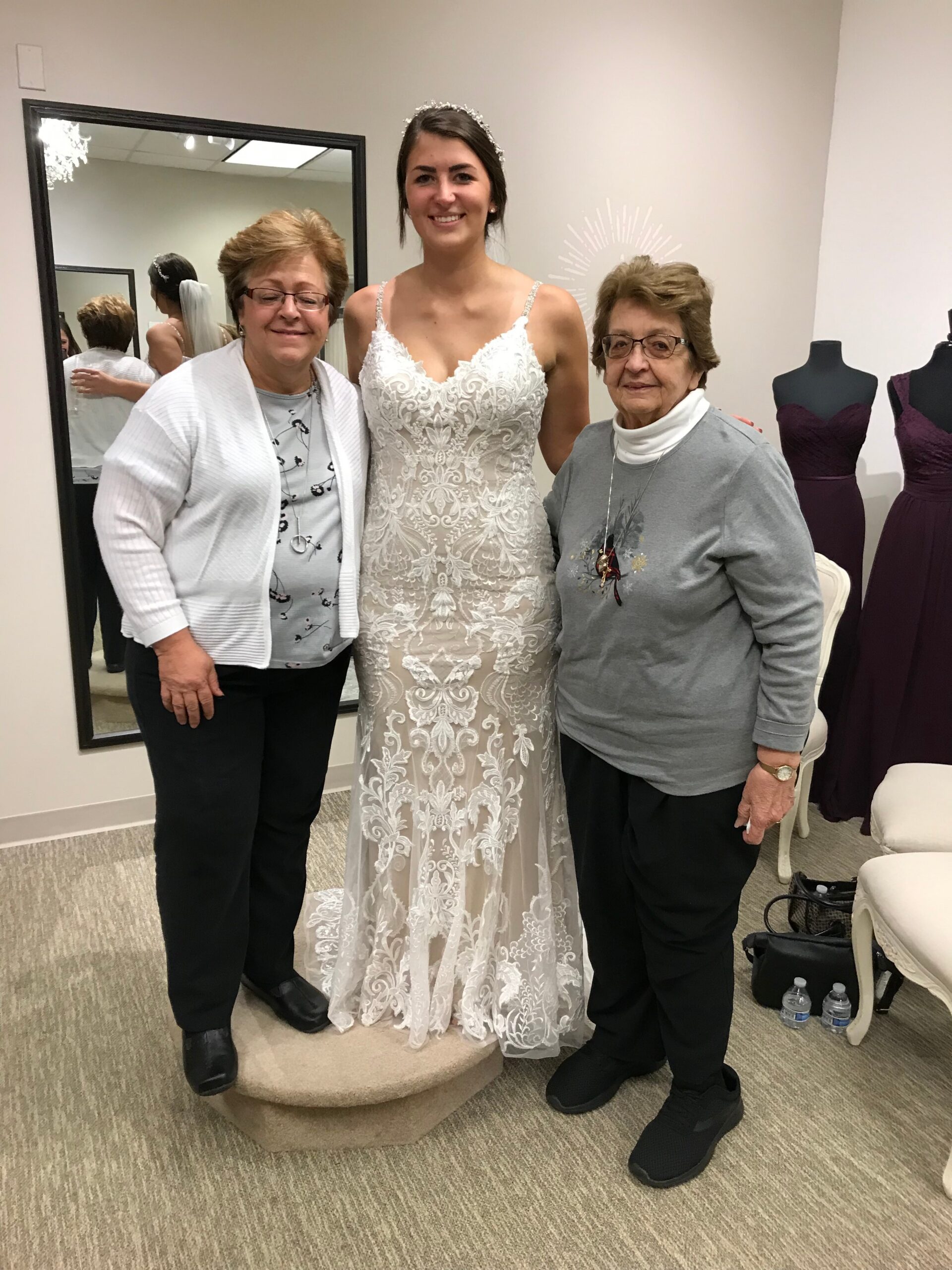 Bride in a white lace wedding gown posing with two women in a bridal shop fitting room.