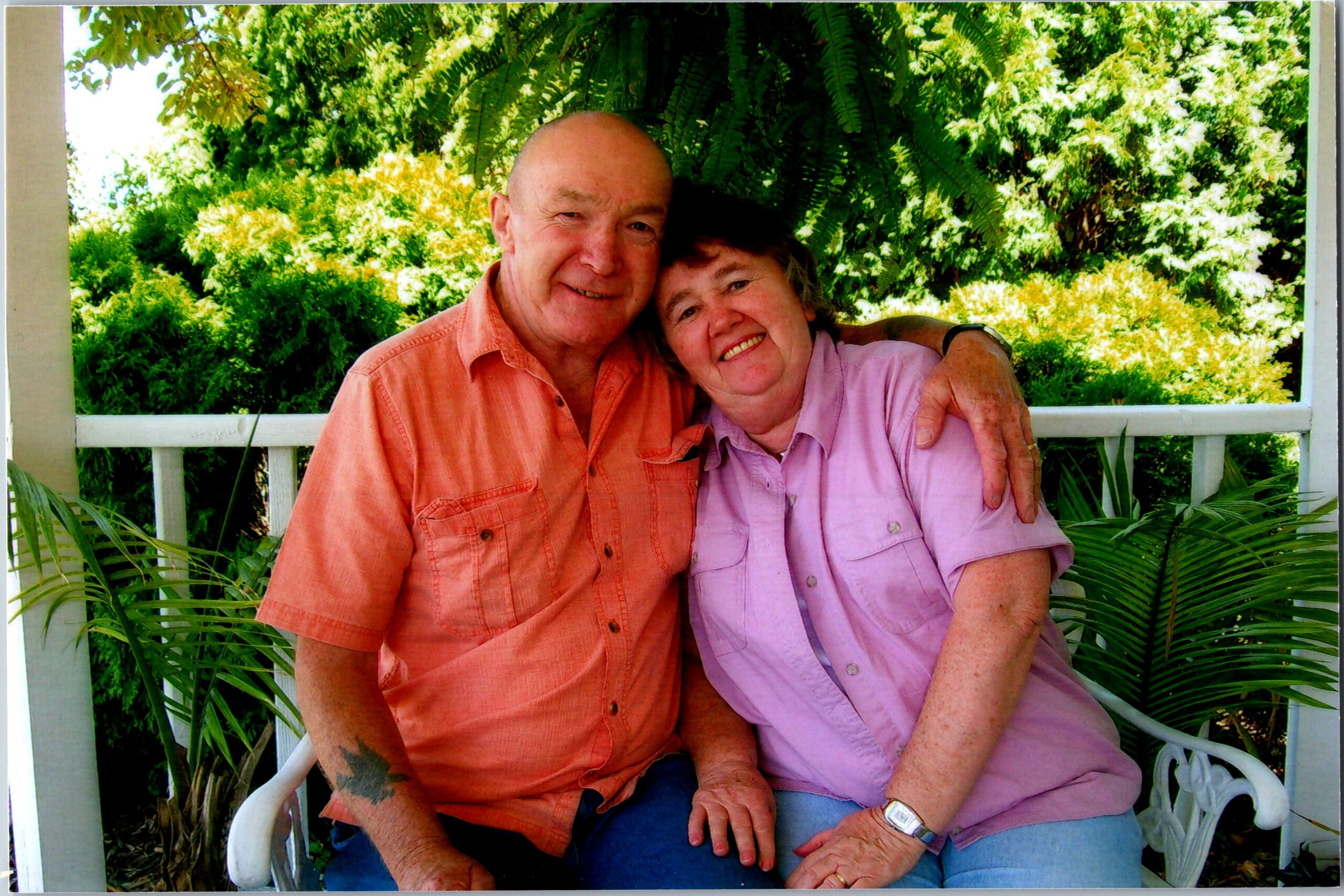 Smiling elderly couple sitting close together on a porch, with lush green plants in the background.