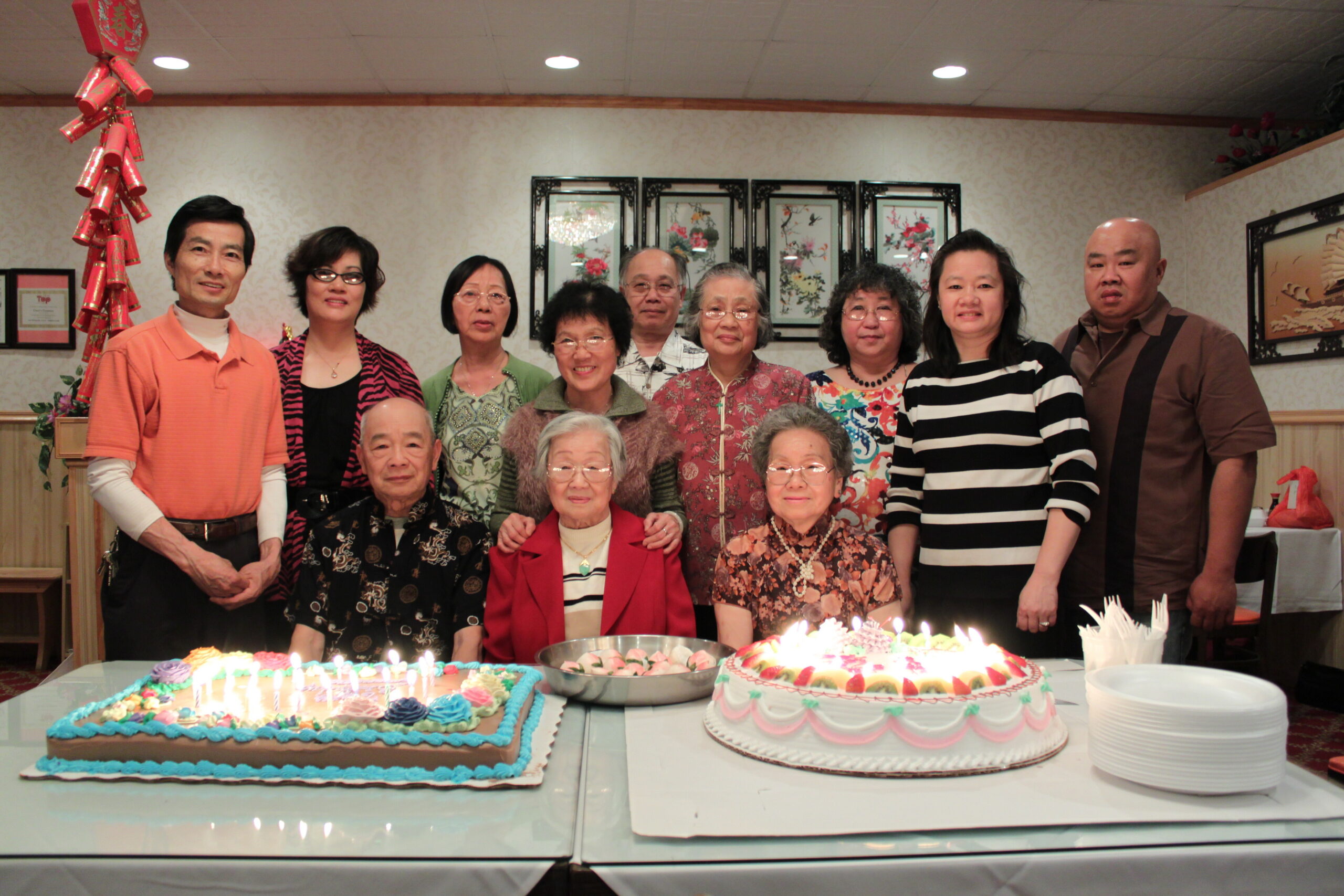 Group of mixed-age adults posing near a long table with two large celebratory cakes, candles lit, in a decorated room.
