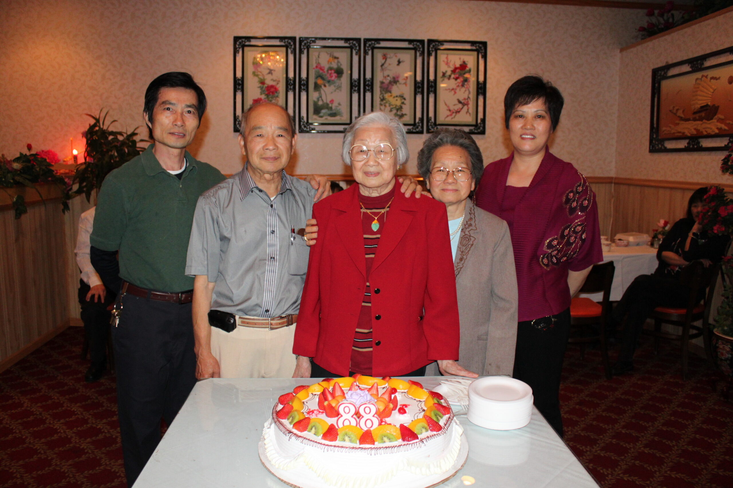 Five adults of Asian descent pose behind a table with a fruit-topped birthday cake displaying the number 88 in a decorated room.
