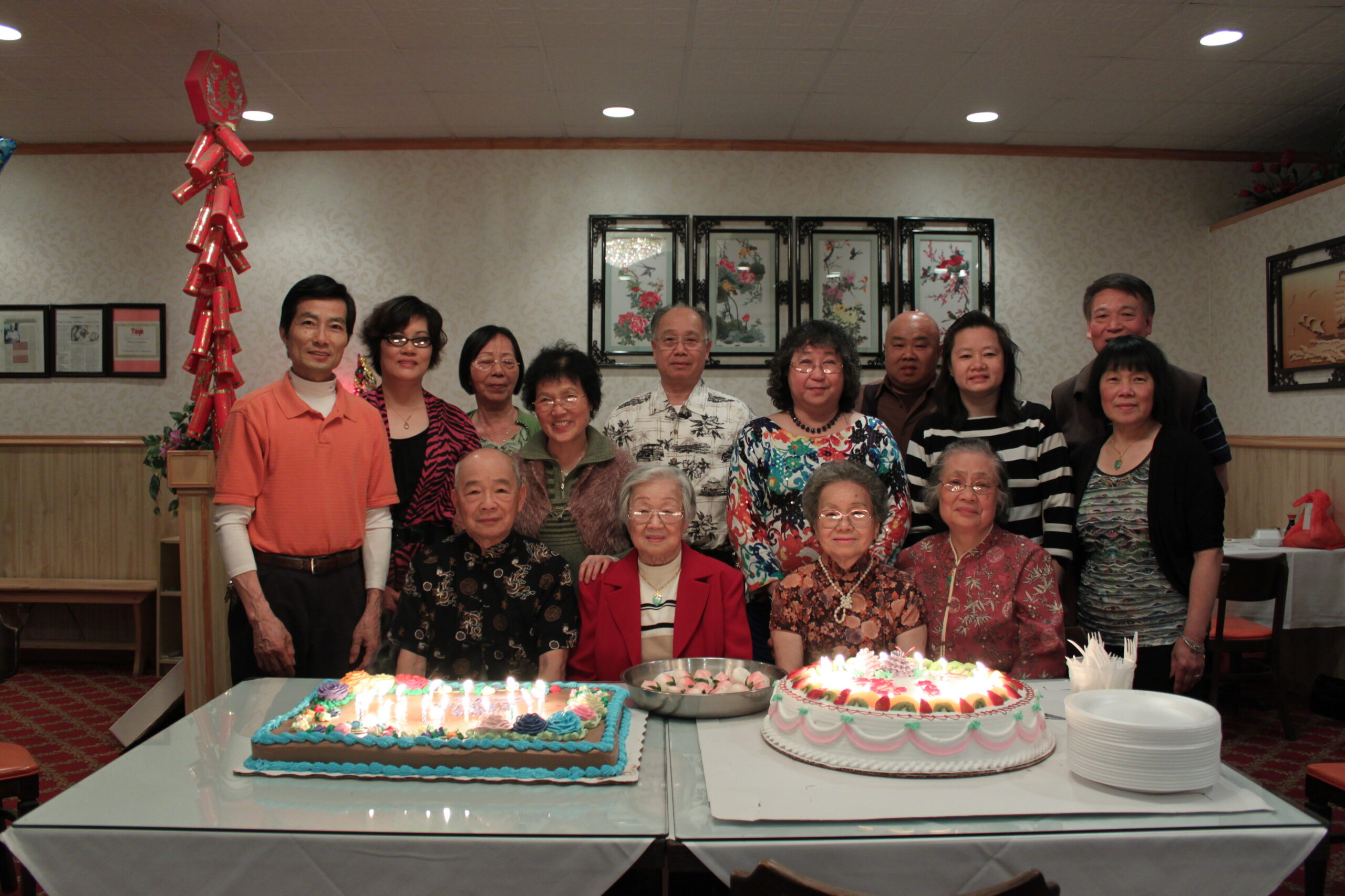 Group of adults posing behind a table with two large birthday cakes and candles, in a party setting.