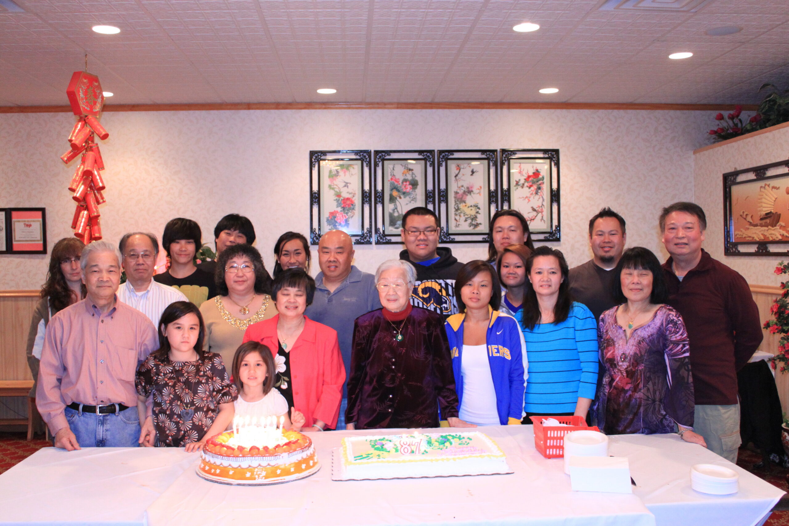 Group of adults and children posing behind a table with two birthday cakes in a banquet room decorated with red Chinese ornaments and framed art on the wall.