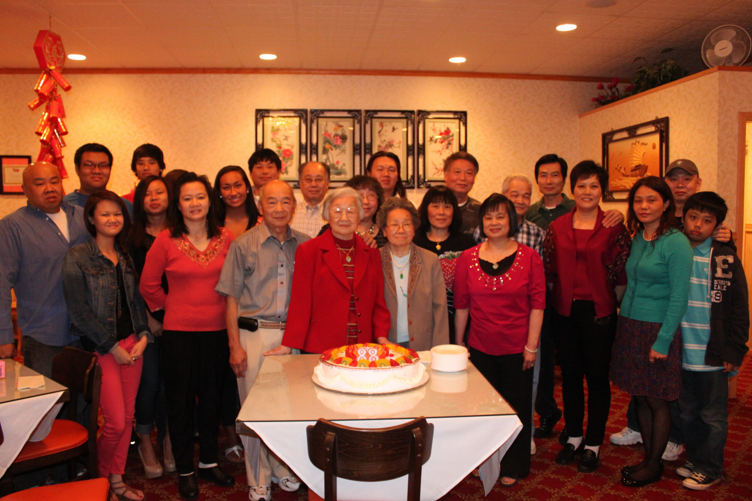 Group of people of various ages posing around a large birthday cake on a dining table inside a restaurant.