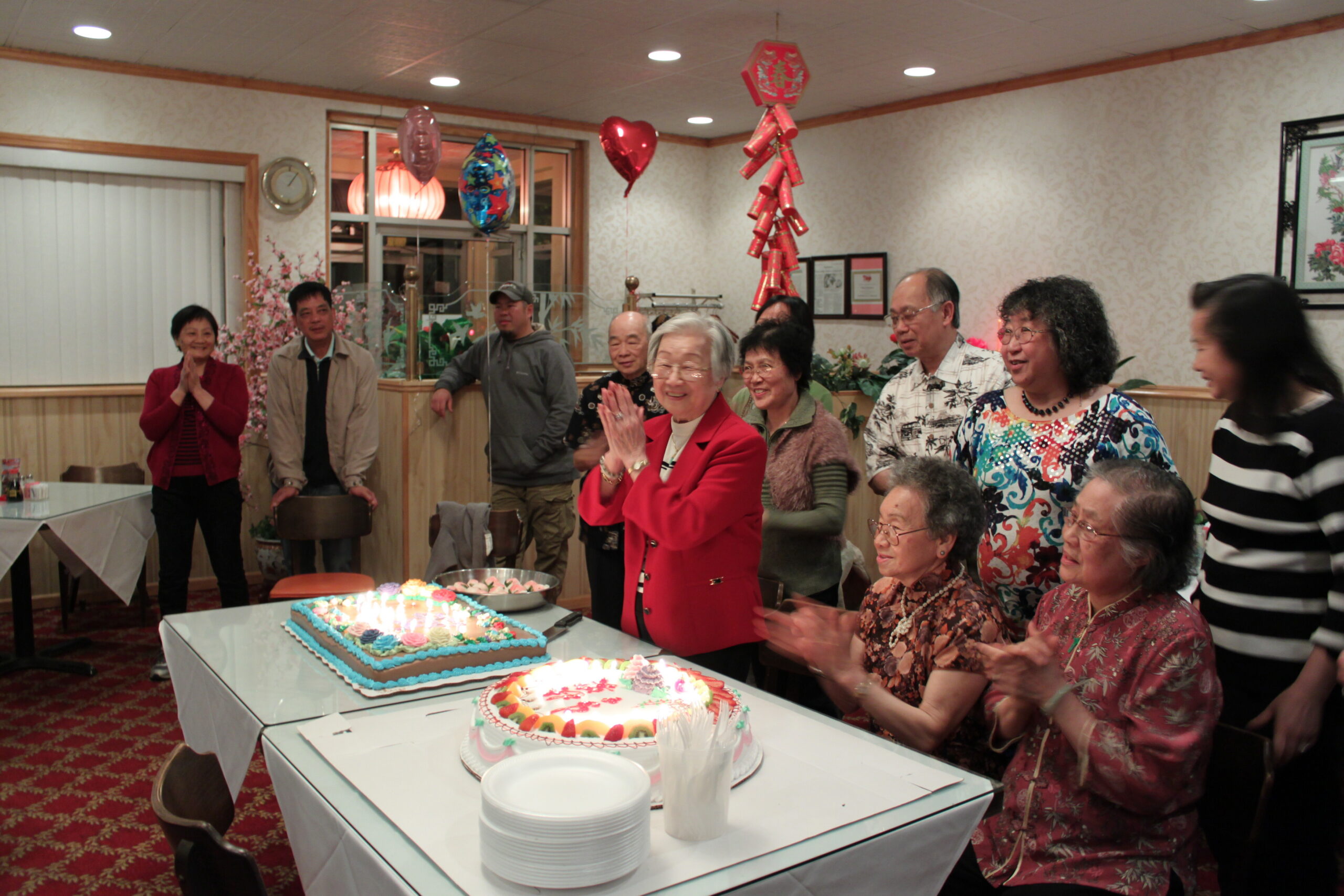 A group of seniors gathered around a table with a large tiered cake and cupcakes for a celebration in a decorated dining room, some clapping and smiling at the center.