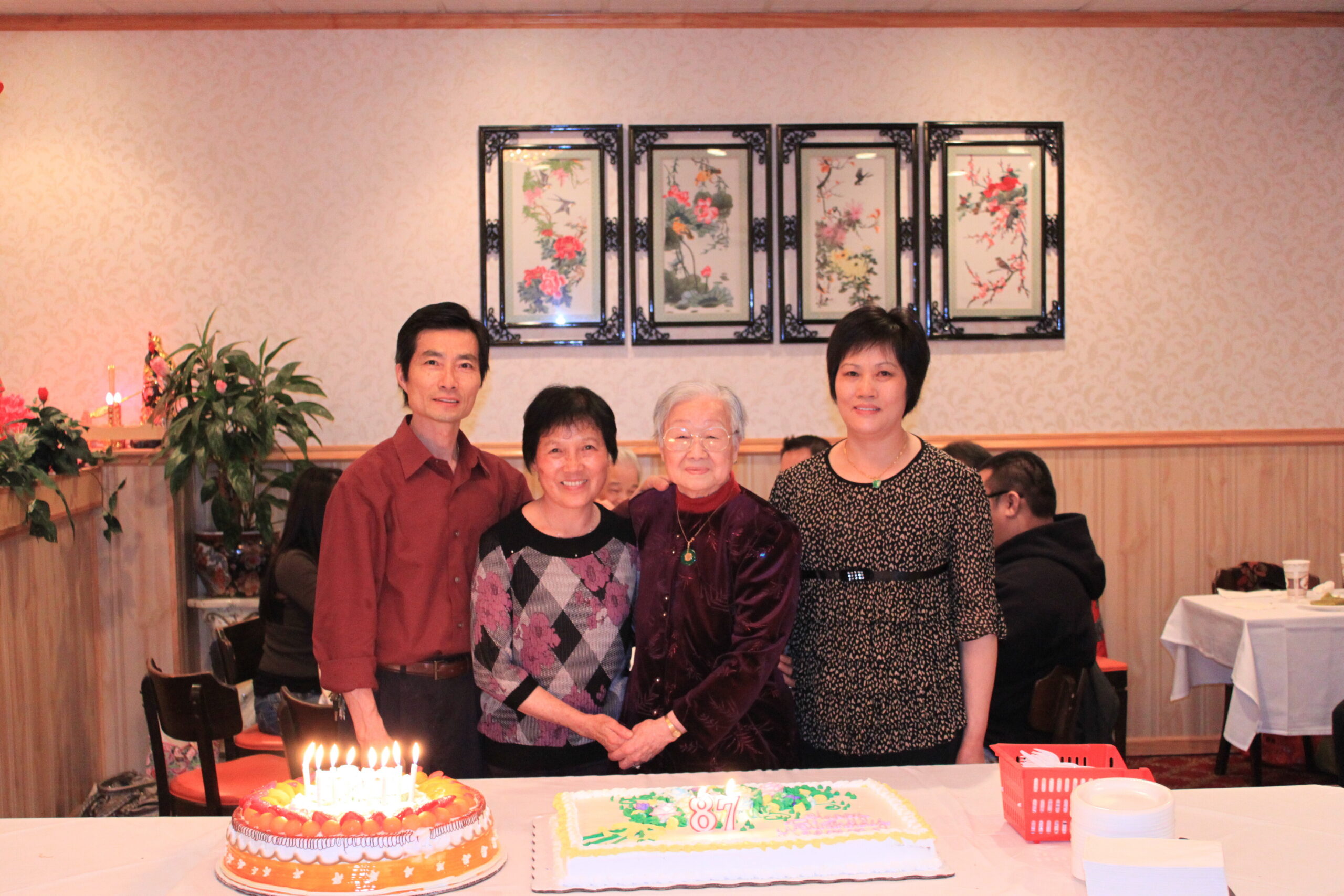 Four people posing for a birthday party photo, with two decorated cakes and candles on the table in front of them.
