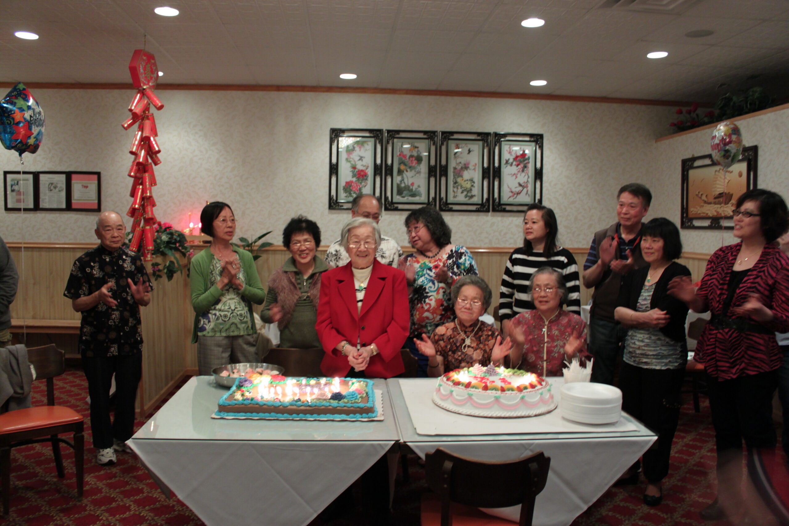 Group of mostly elderly people standing around a table with two large decorated birthday cakes, celebrating in a banquet room with balloons and decorative screens on the wall.